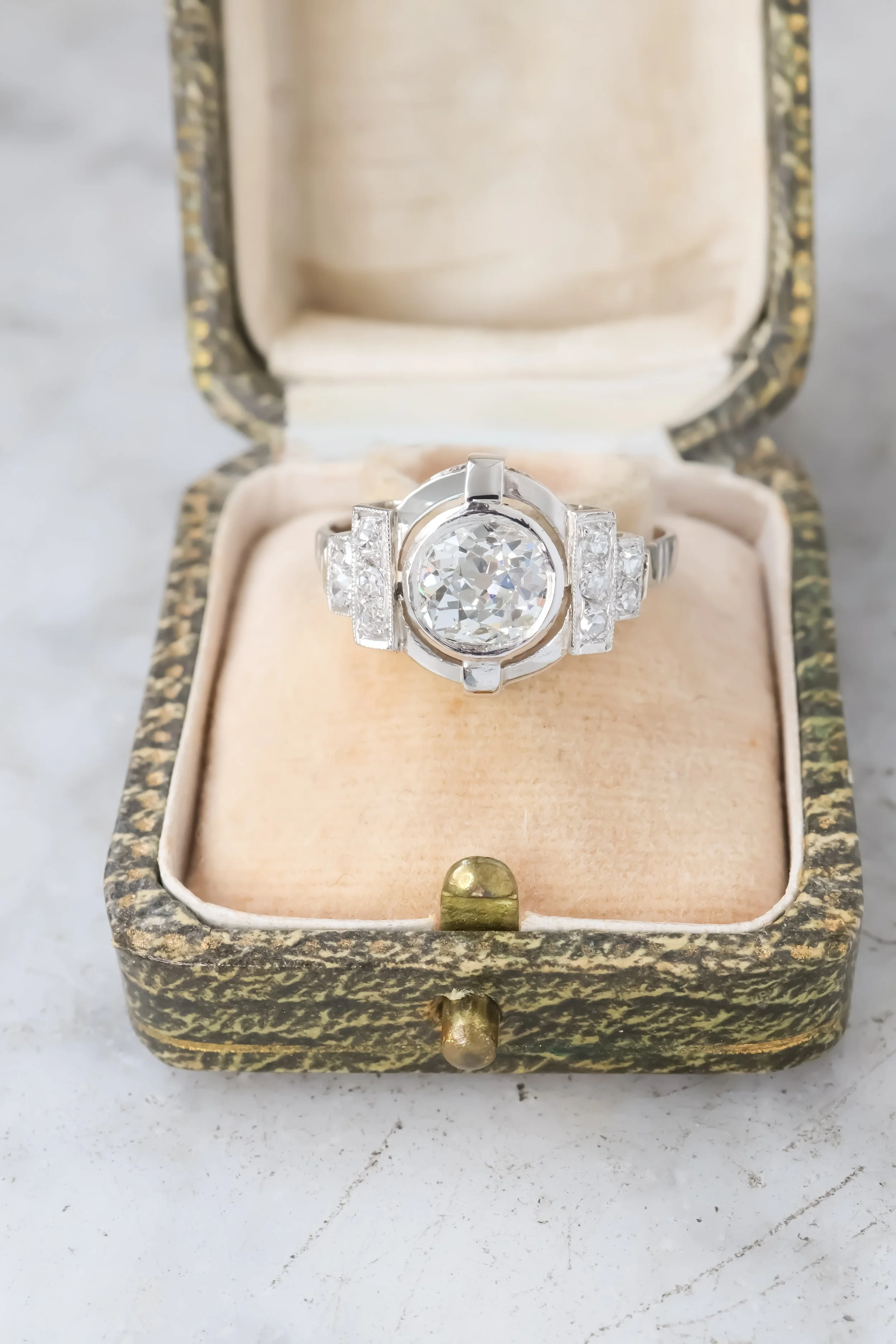 Bague en argent avec un grand diamant rond au centre, accompagnée de diamants plus petits de chaque côté, dans une boîte à bijoux ancienne ornée de motifs dorés.