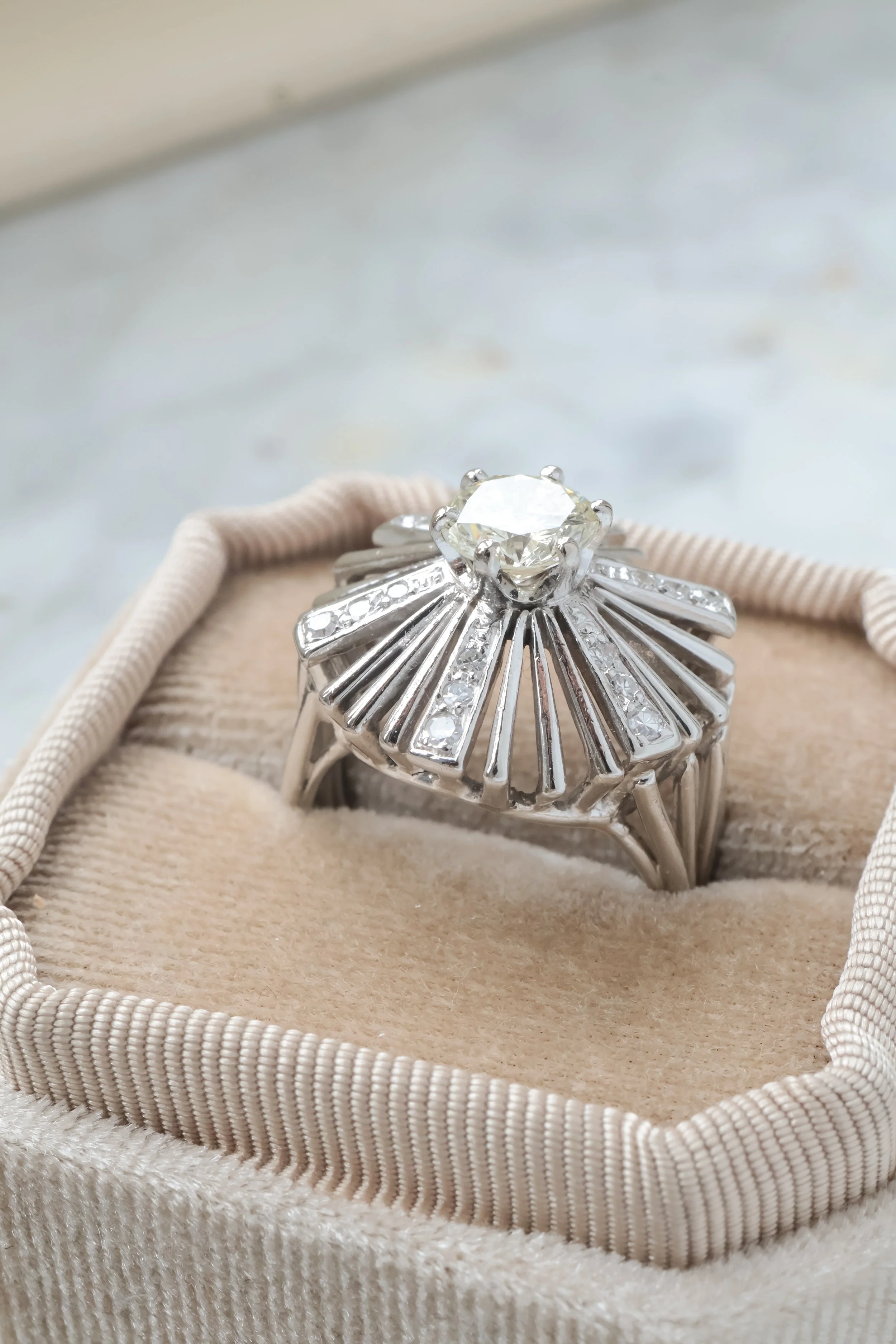 Une bague en argent ornée d'un gros diamant central, posée dans une boîte en velours beige.