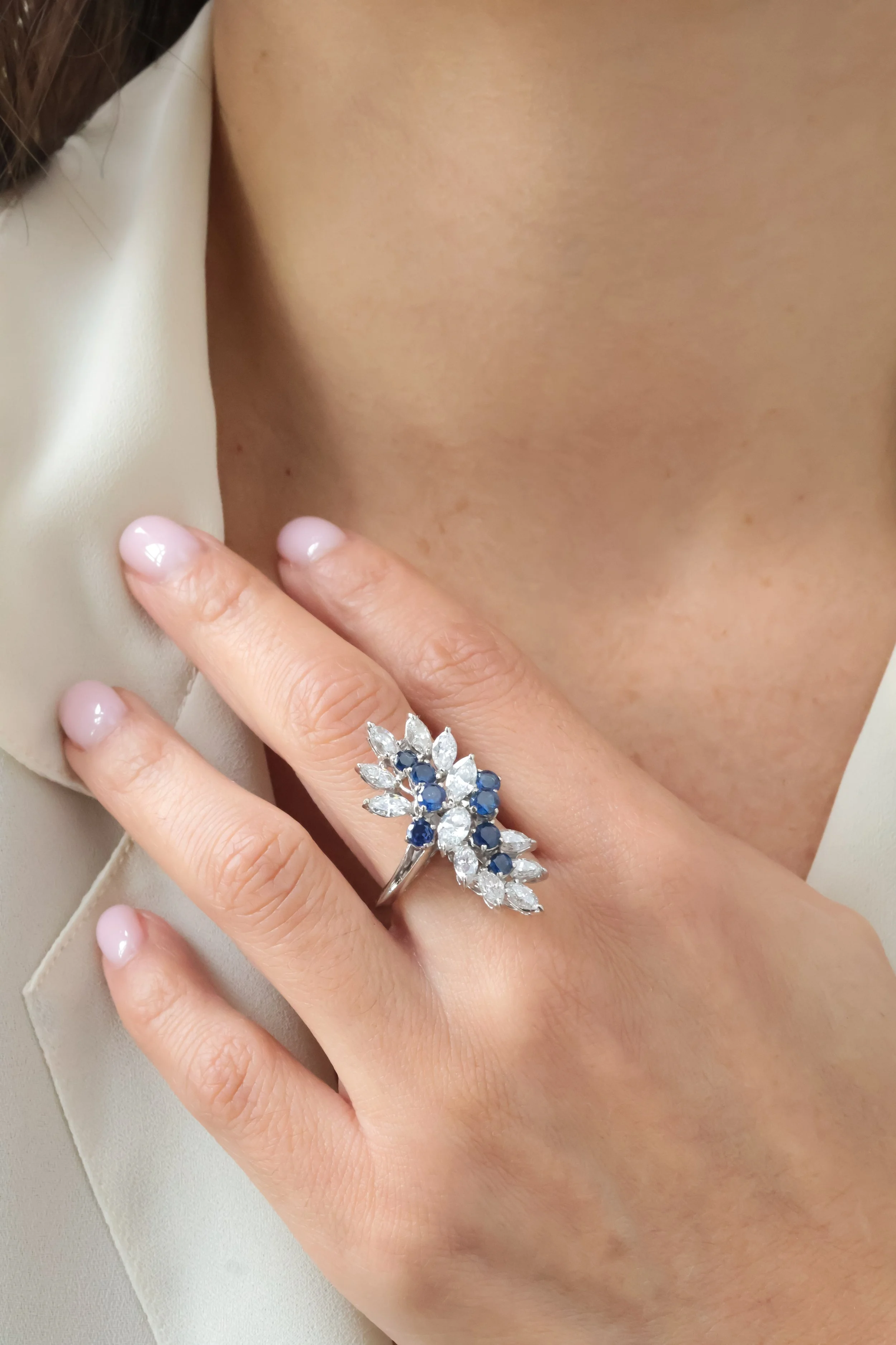 Bague cocktail en or blanc ornée de diamants taille navette et de saphirs ronds dans un écrin beige, portée à la main en zoomant 