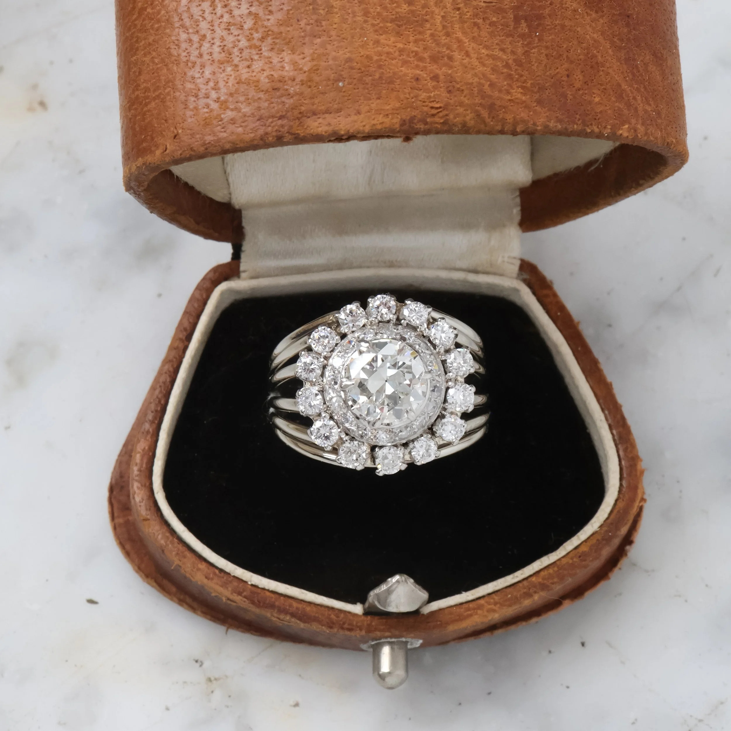 Bague en argent avec un grand diamant central entouré de petits diamants, présentée dans une boîte en bois marron avec intérieur en tissu blanc et noir.