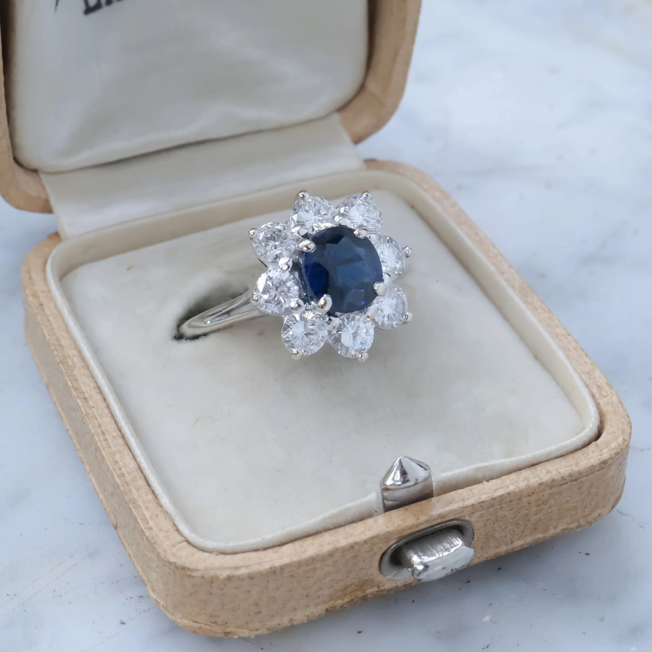 Une bague en argent avec un saphir bleu entouré de diamants, dans une boîte à joyaux en velours beige.