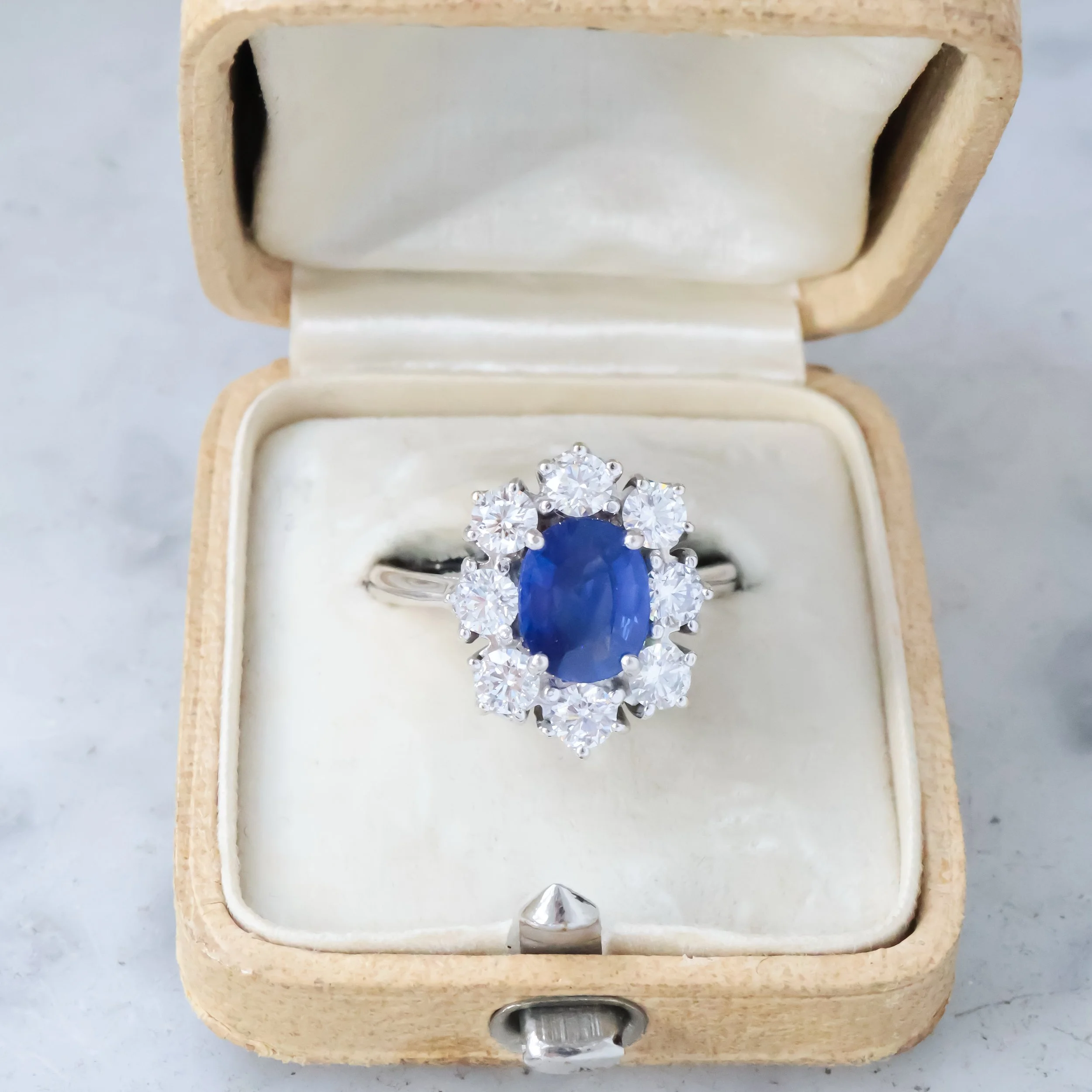 Bague en or avec un saphir ovale au centre entouré de diamants, dans un écrin à bijou vintage avec intérieur en satin blanc.