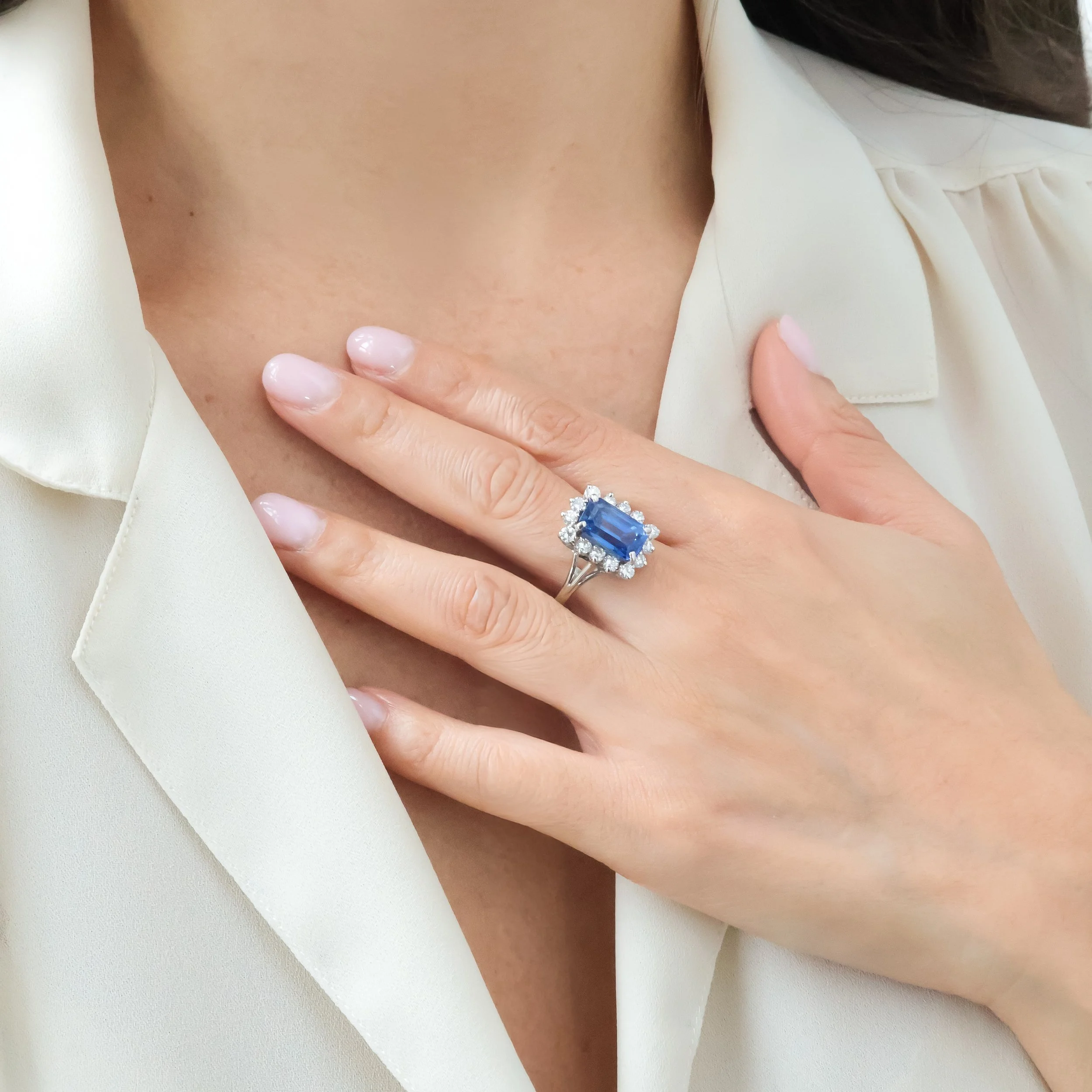 Une main portant une bague avec un grand diamant bleu entouré de petits diamants, posée sur la poitrine.