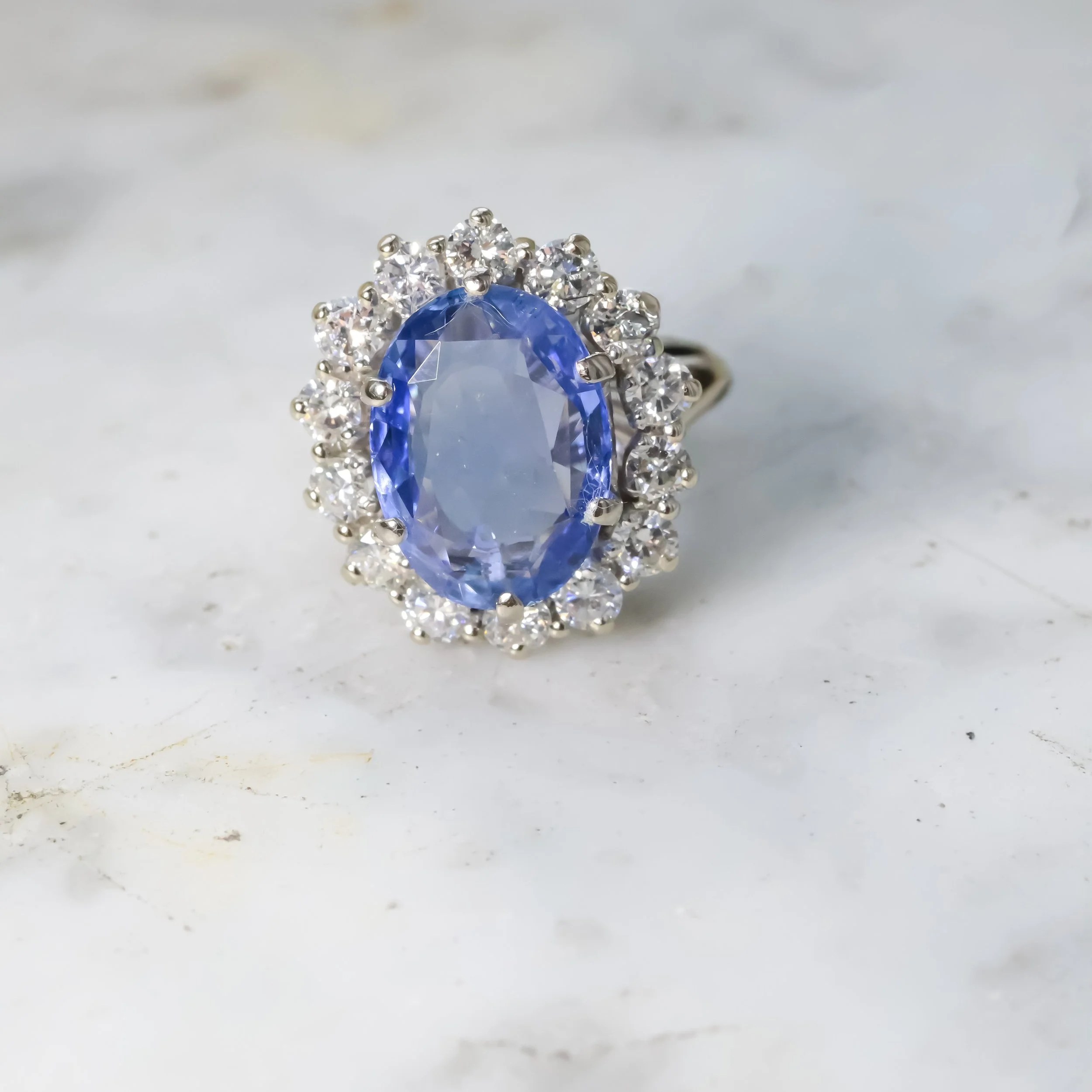 Bague avec un grand pierre ovale bleue entourée de petites pierres blanches sur une surface blanche.