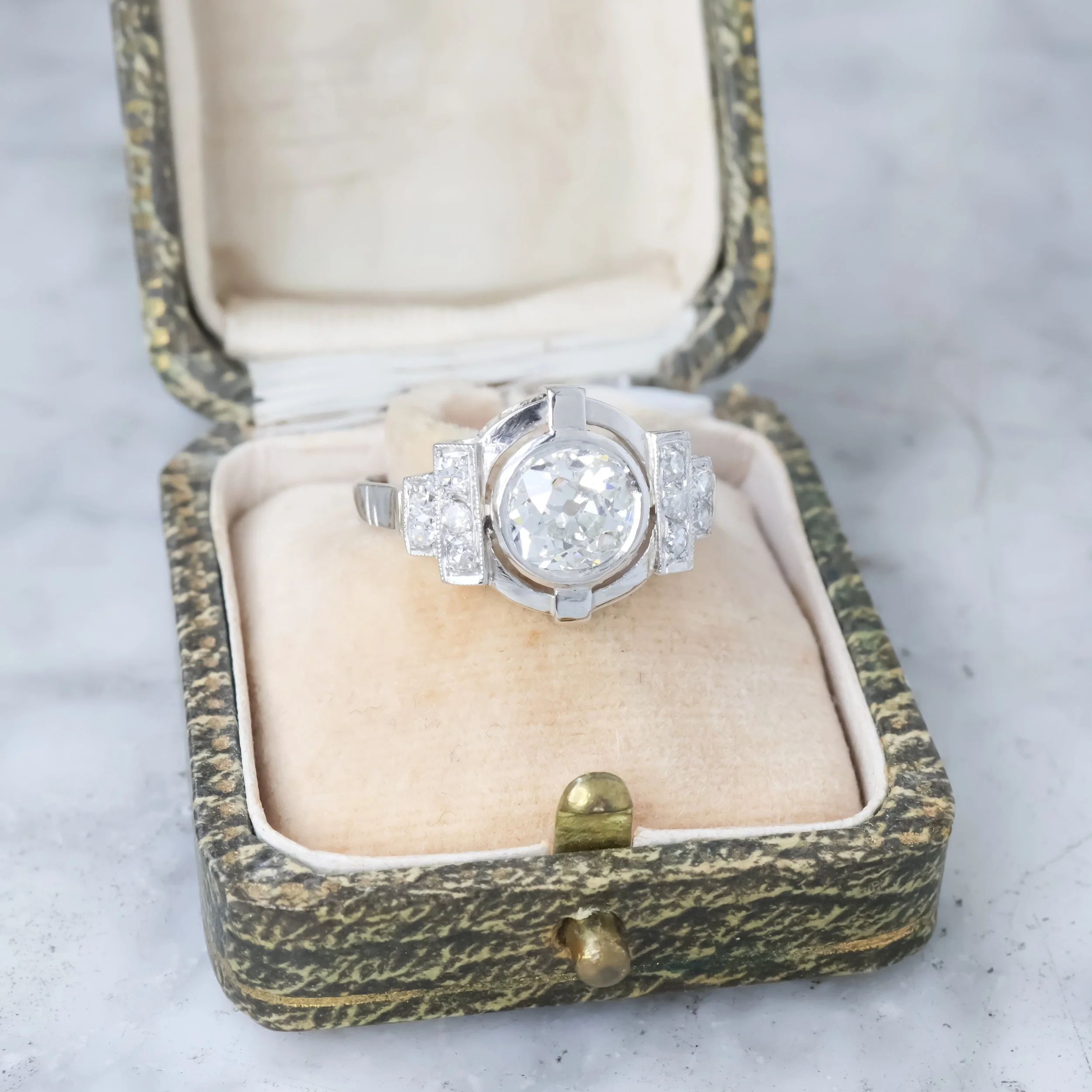 Une bague en platine avec un grand diamant au centre, entourée de petits diamants, dans une boîte à bijoux vintage avec intérieur en velours beige.