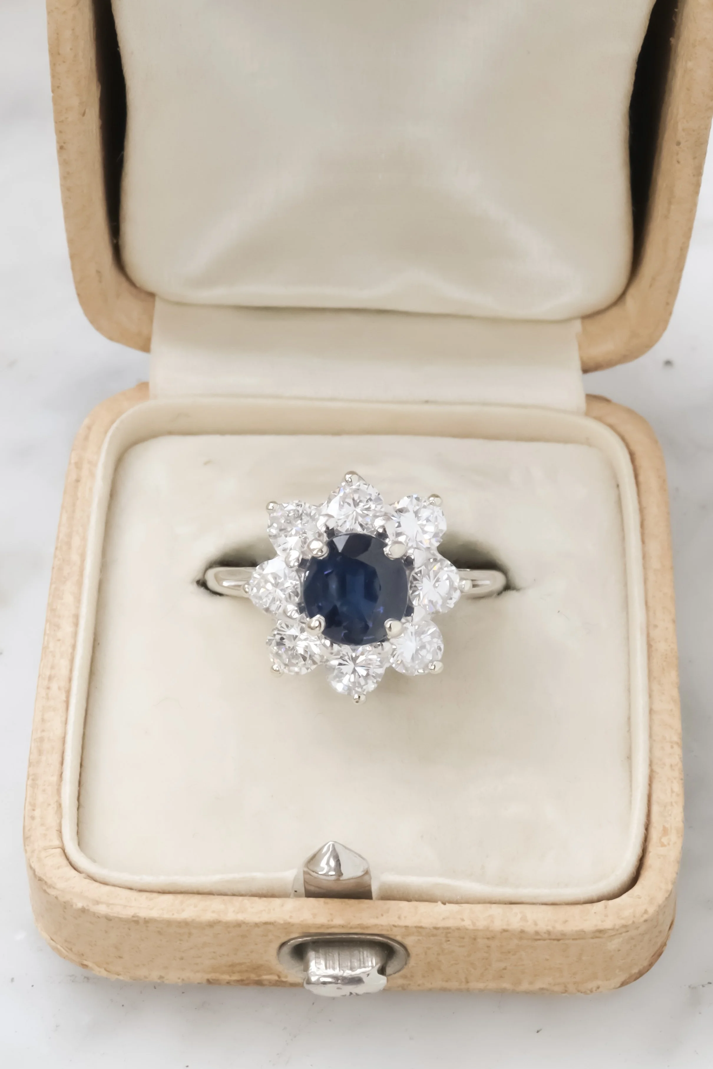 Bague élégante avec un saphir ovalée bleu foncé entouré de diamants, dans une boîte à bijou en velours blanc.