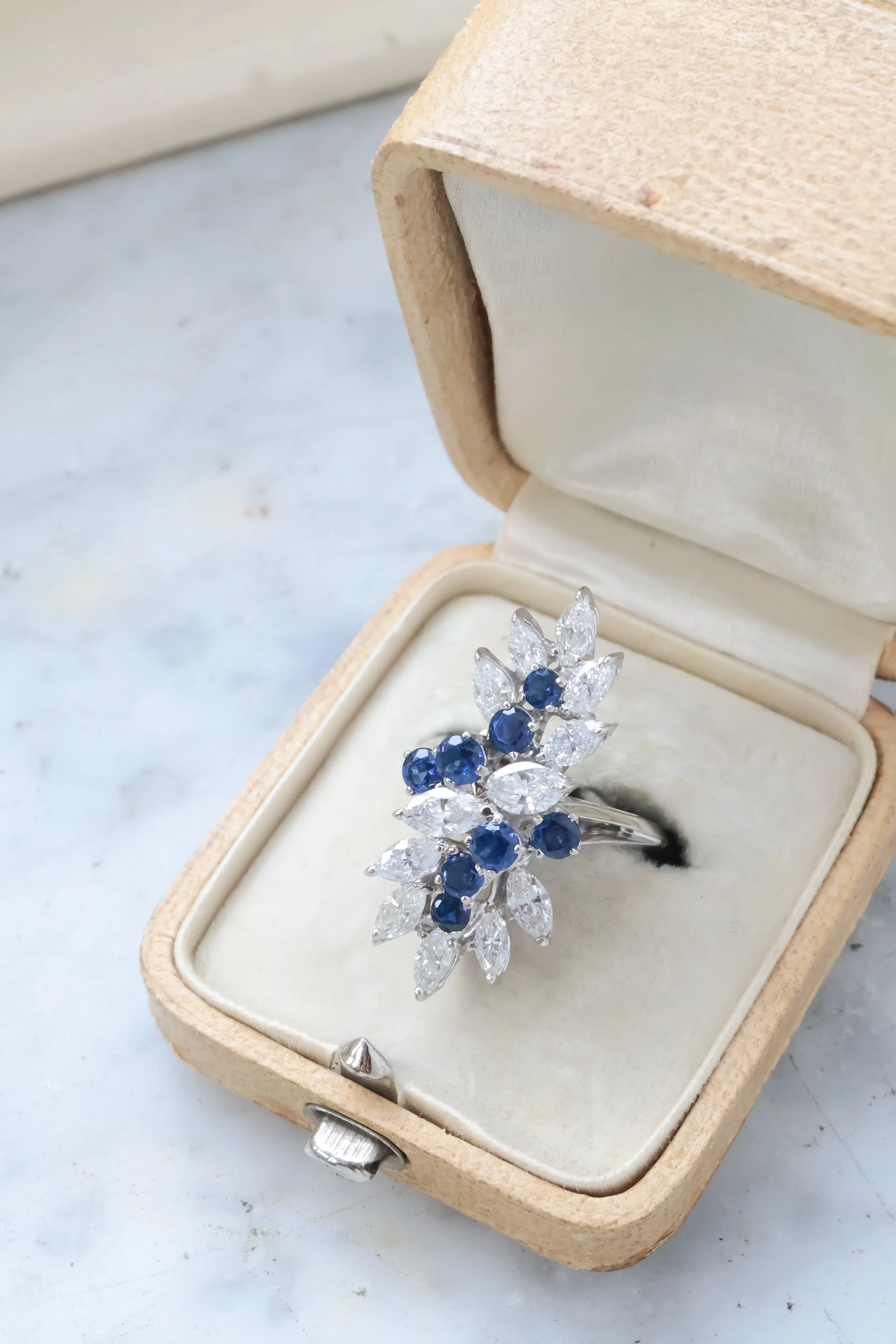 Bague cocktail en or blanc ornée de diamants taille navette et de saphirs ronds dans un écrin beige , vue de côté