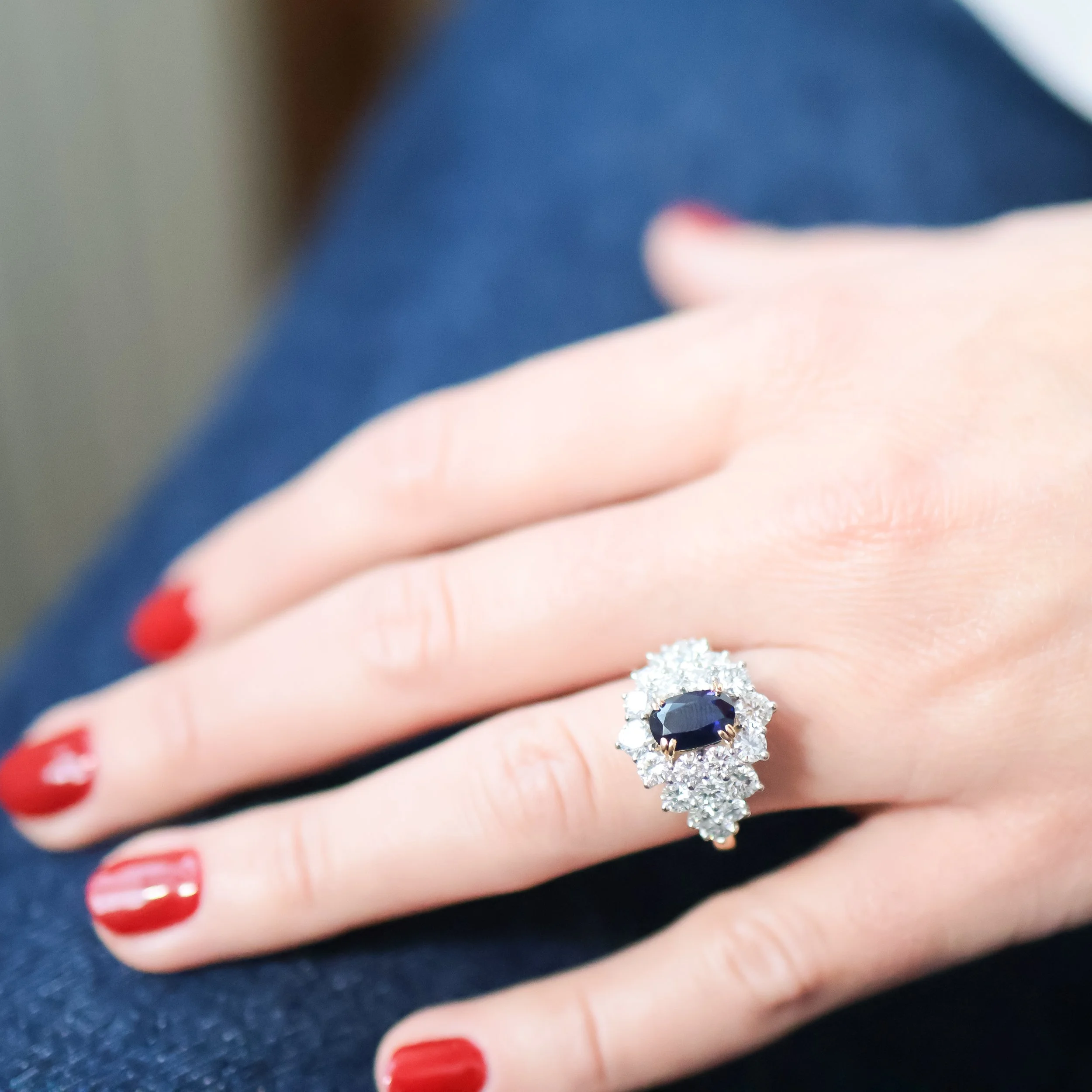 Une main portant une bague ornée d'un gros saphir bleu entouré de diamants, avec des ongles rouge