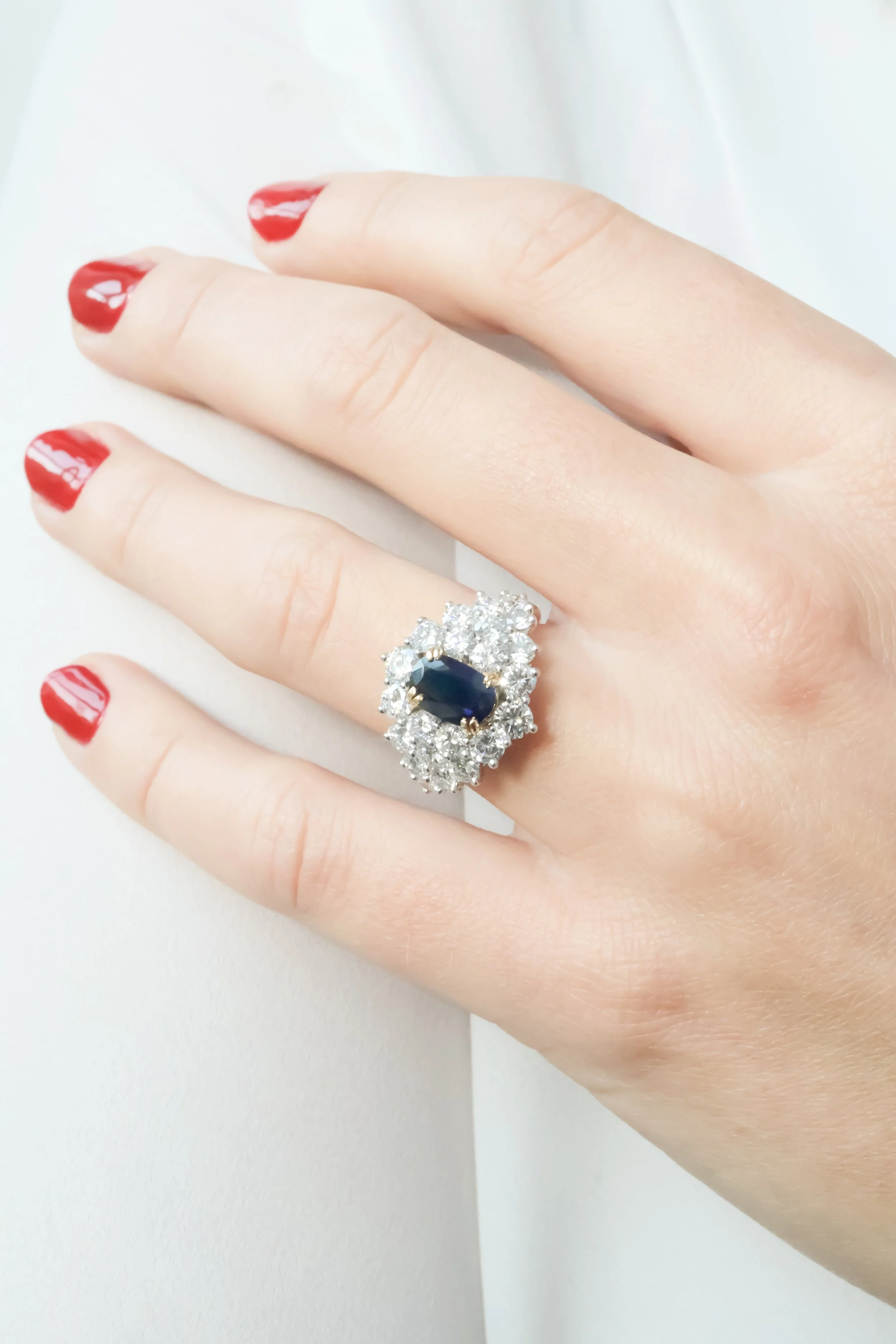 Une main portant une bague ornée d'une pierre centrale sombre entourée de diamants, avec des ongles peints en rouge