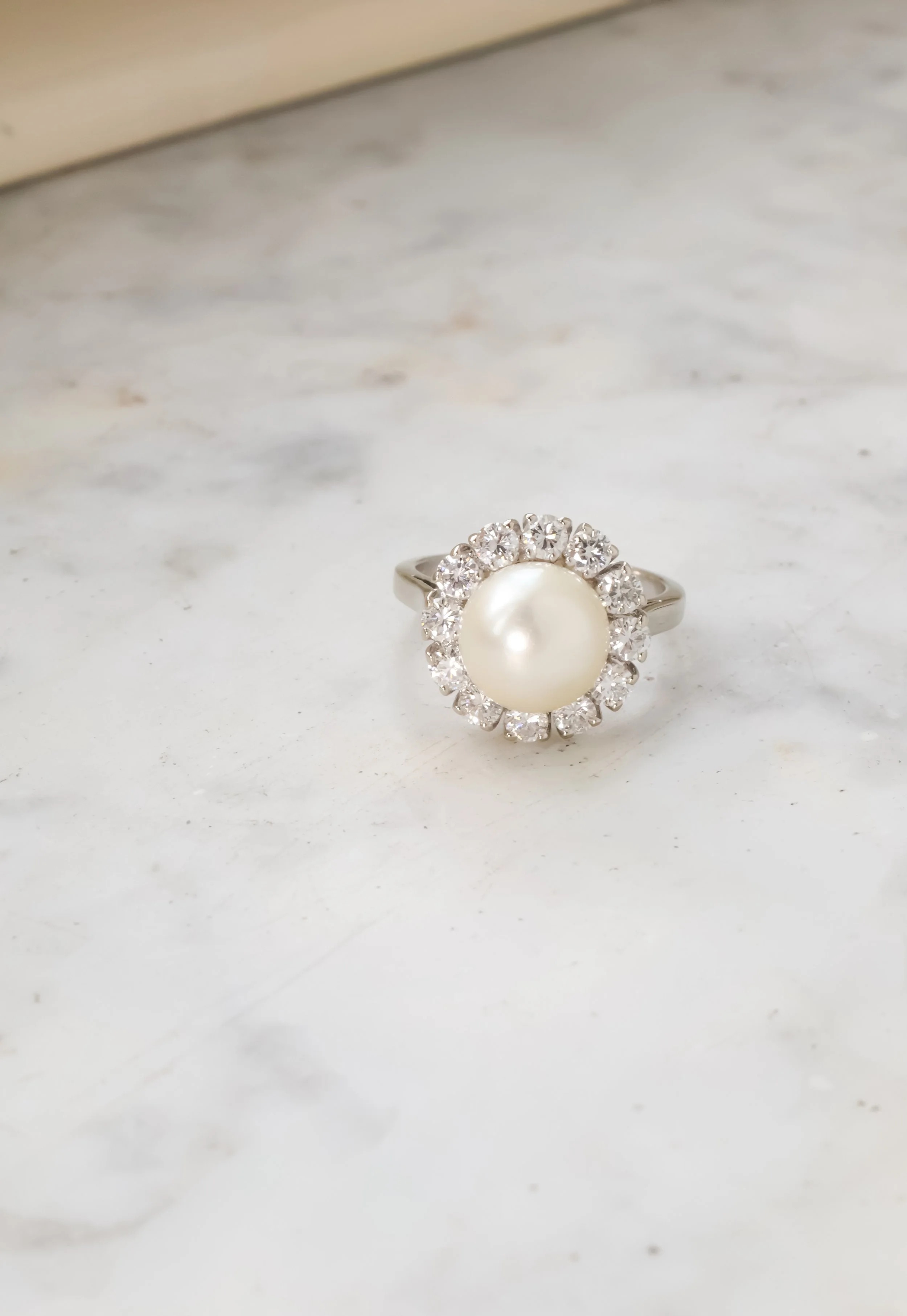 Bague en argent avec une grande perle au centre, entourée de petits diamants, posée sur une surface de marbre.