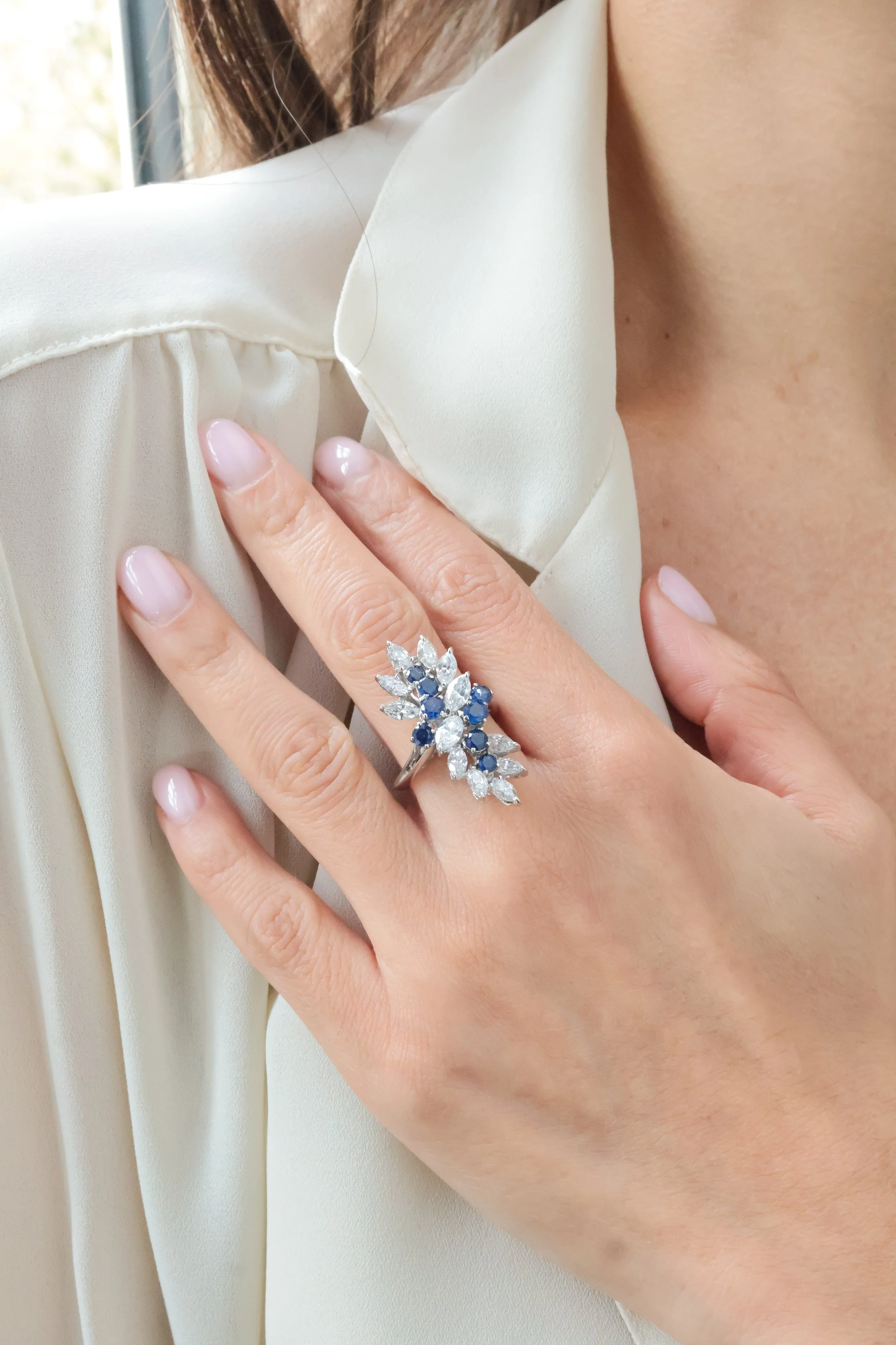 Bague cocktail en or blanc ornée de diamants taille navette et de saphirs ronds dans un écrin beige, portée à la main 