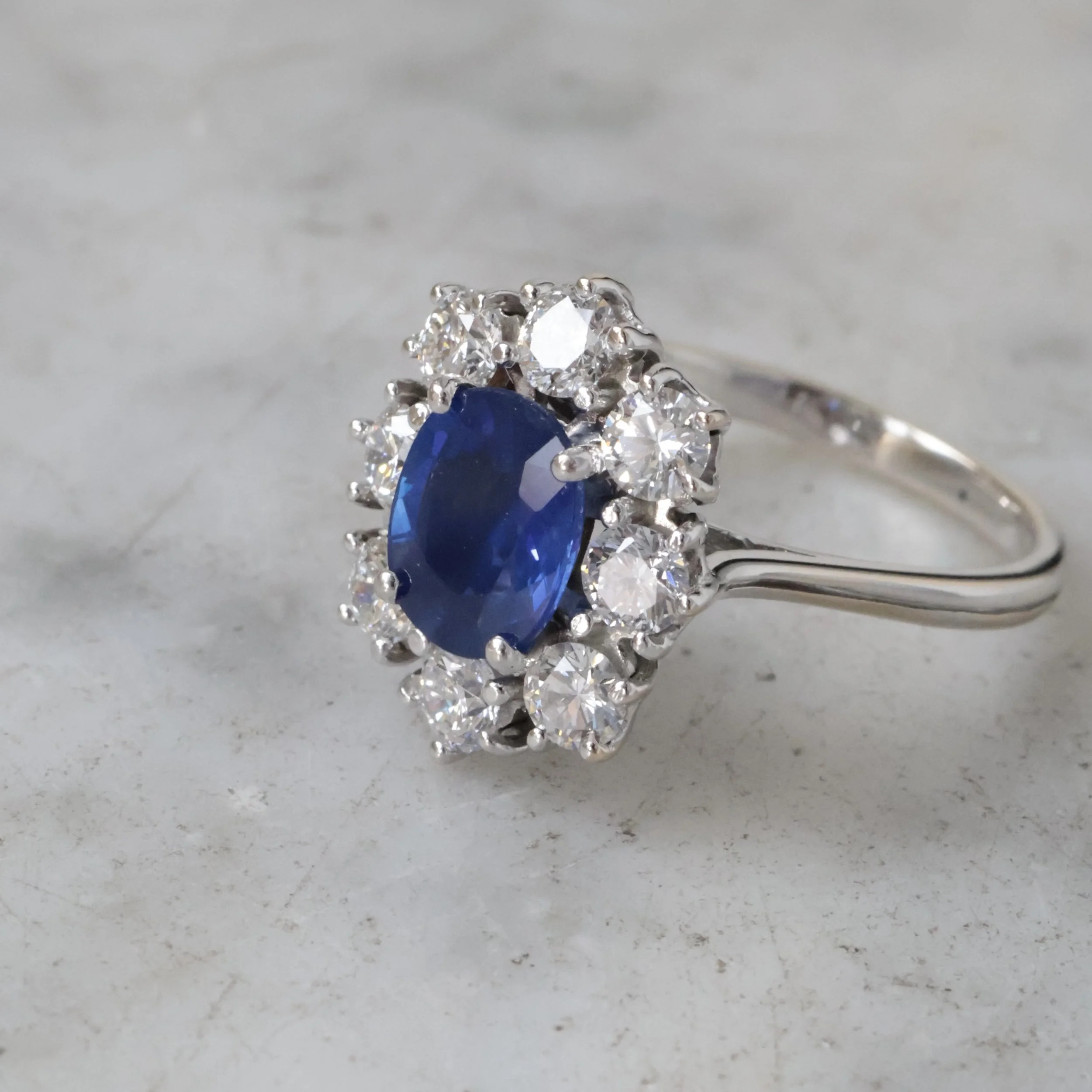 Bague ancienne en or blanc avec saphir ovale bleu entouré de diamants, posée sur fond clair