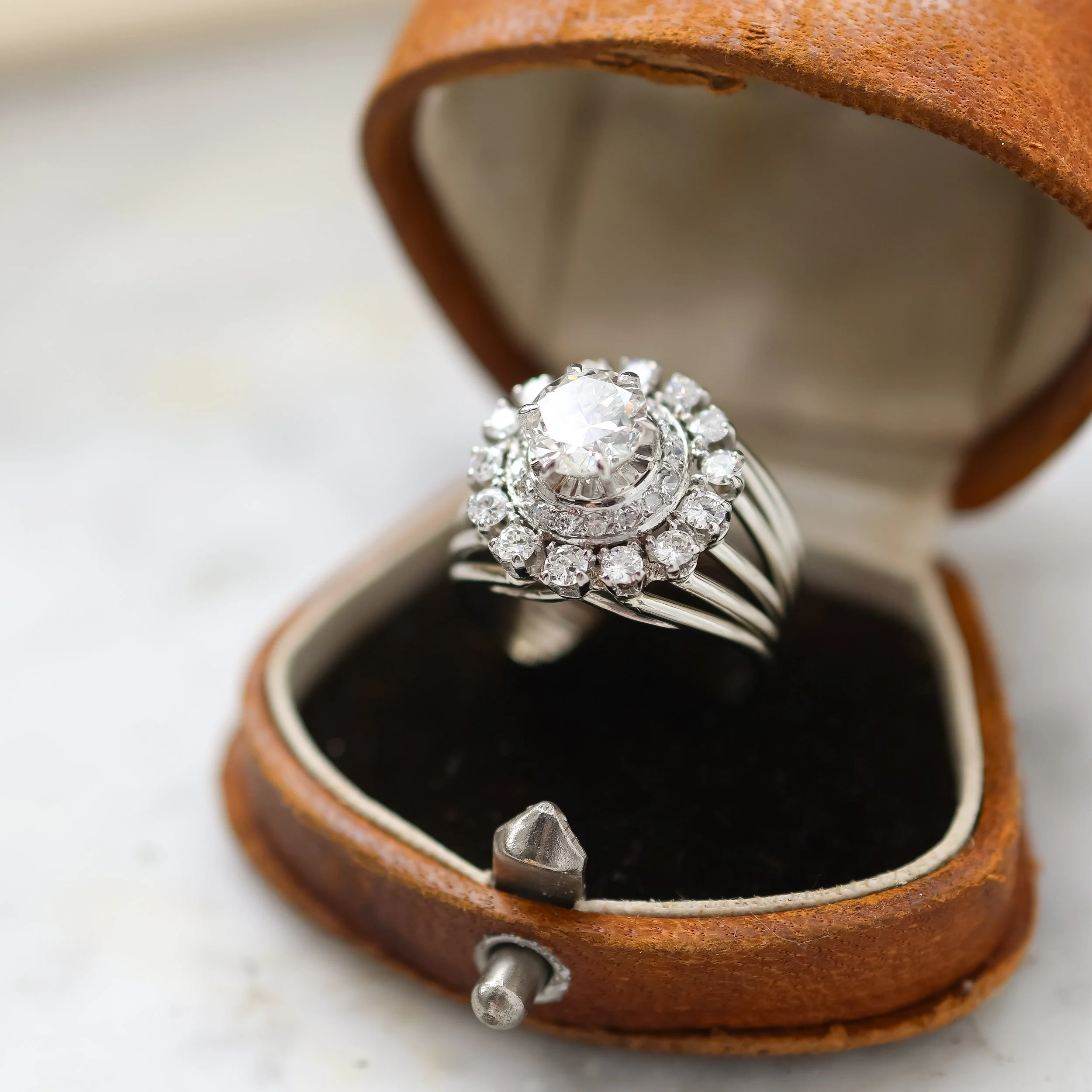 Une bague en argent avec un grand diamant au centre entouré de petits diamants dans une boîte en cuir marron.