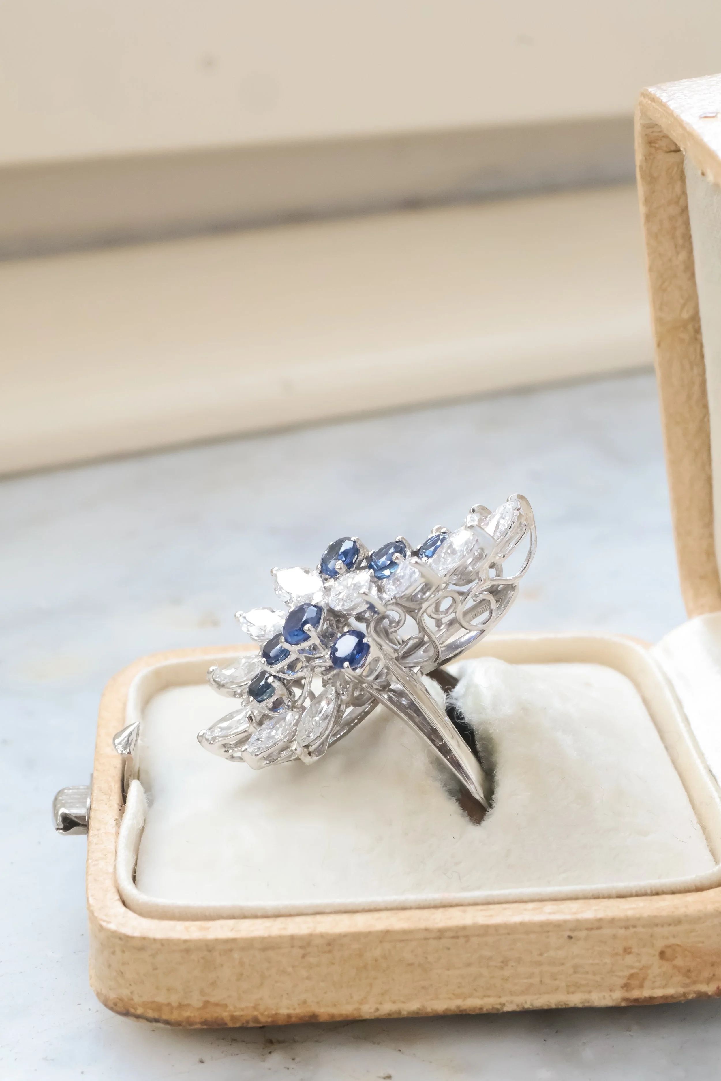 Bague cocktail en or blanc ornée de diamants taille navette et de saphirs ronds dans un écrin beige, vue de profil