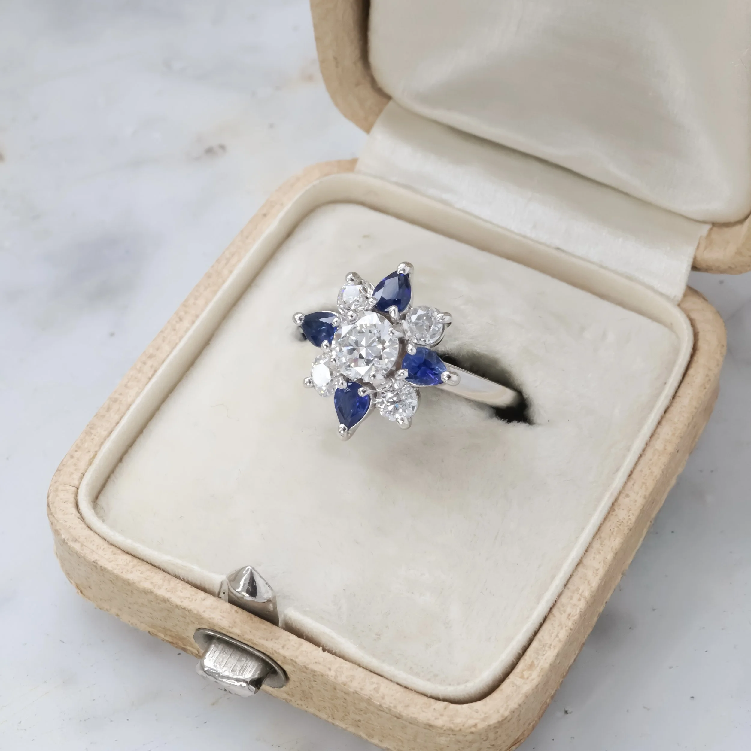 Bague en or blanc ornée de diamants et de saphirs bleus dans une boîte à bijoux vintage en velours blanc.
