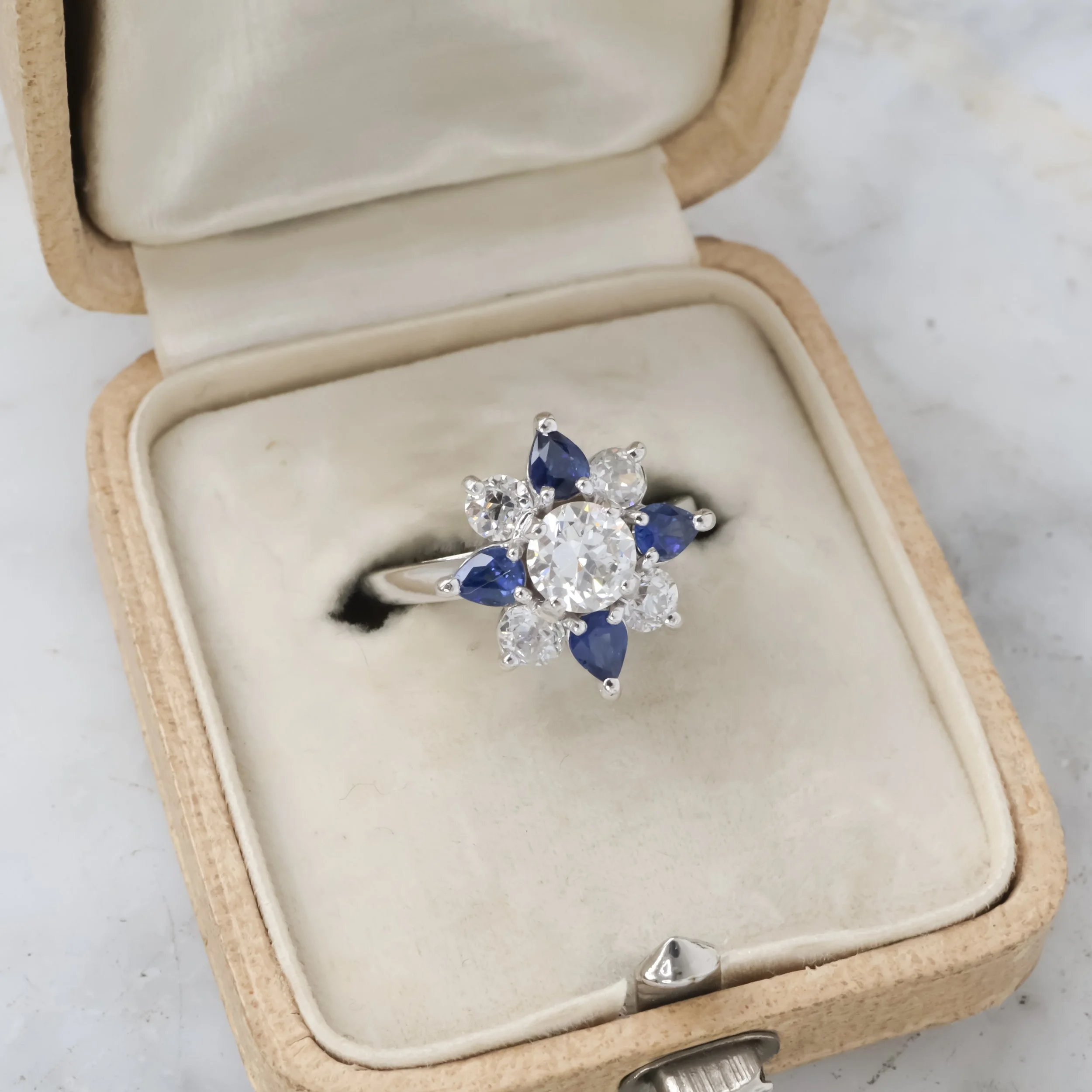 Bague en or blanc avec un centre en diamant entouré de pierres précieuses en forme de fleurs, composées de diamants et de saphirs bleus, présentée dans un écrin vintage beige 