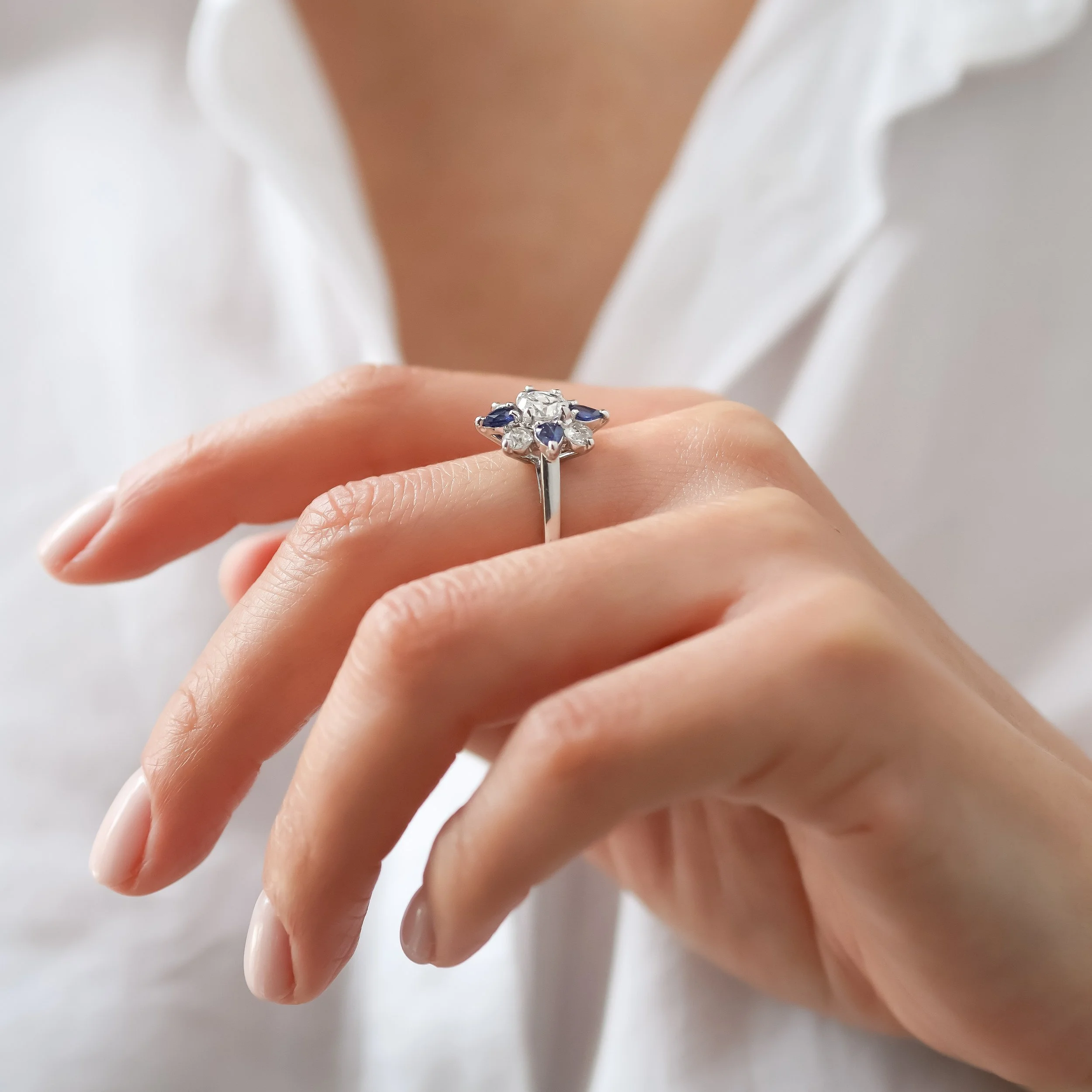 Une main portant une bague en or blanc ornée d'un grand diamant central entouré de petits diamants et de quelques saphirs taille poire, avec un fond flou de vêtement blanc.