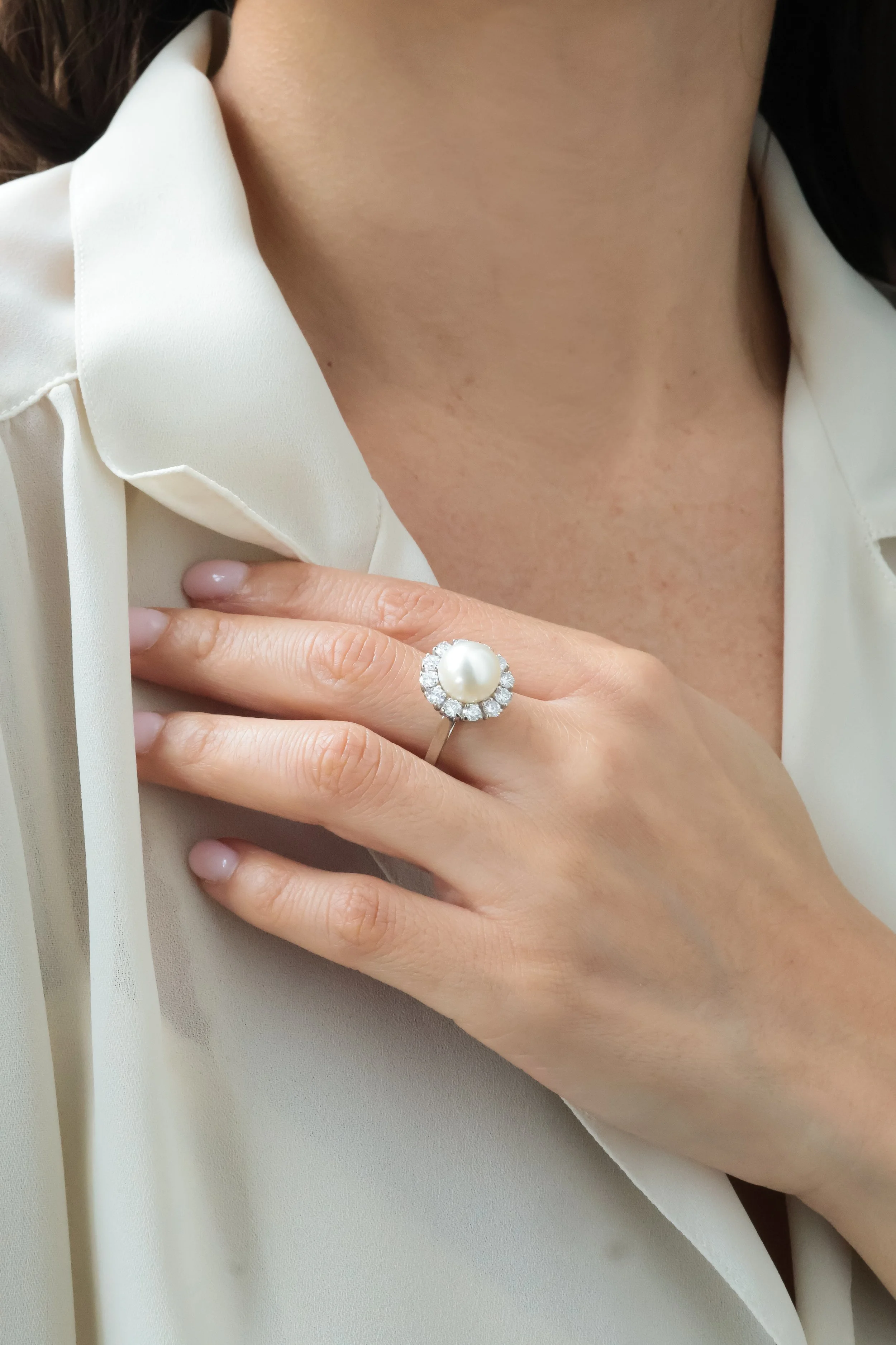 Une main portant une bague avec une pierre centrale en perle entourée de petites pierres brillantes, sur une peau nue près du cou, avec une chemise blanche