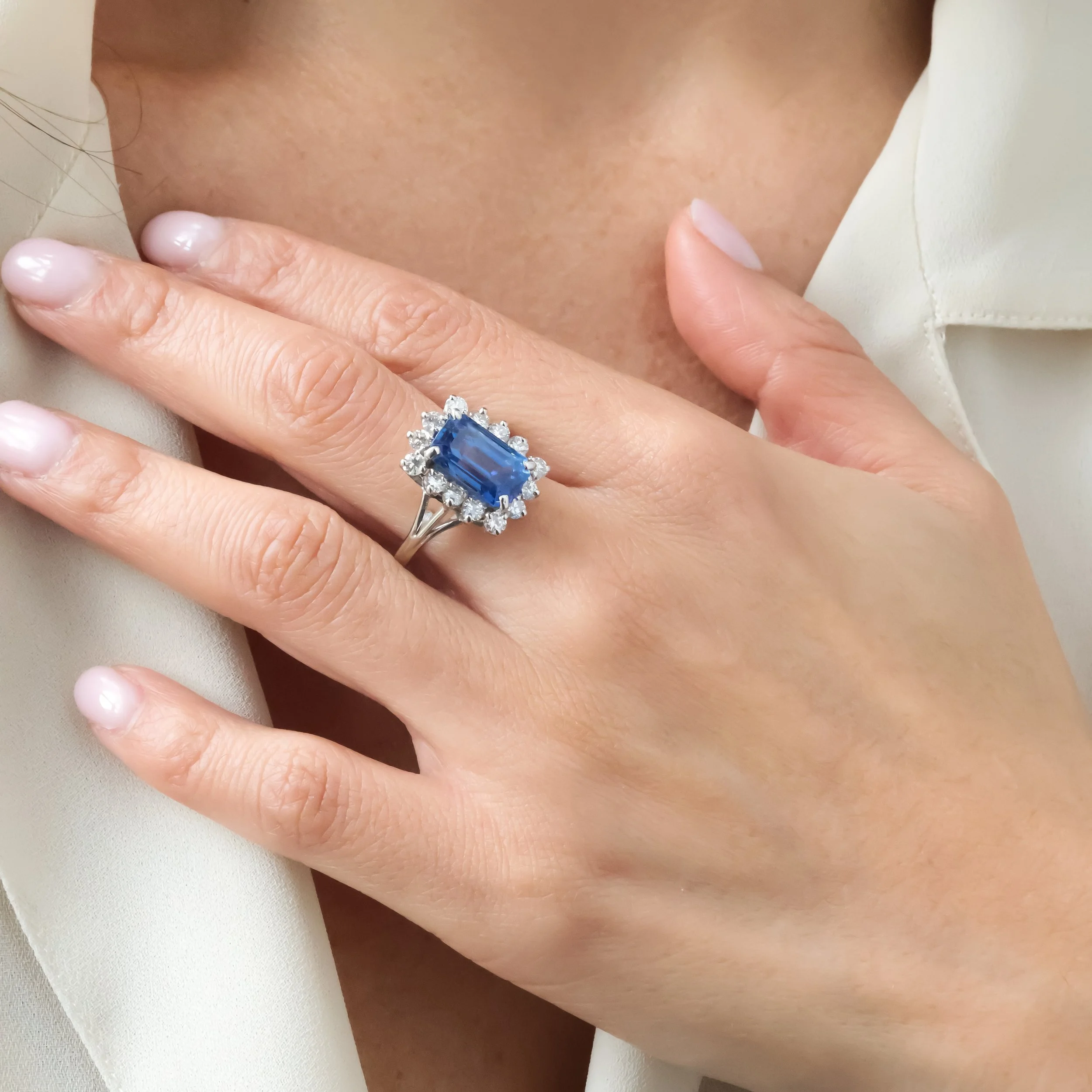 Doigt portant une bague avec un grand saphir bleu entouré de diamants, posé sur la peau d'une personne avec un vêtement beige clair.