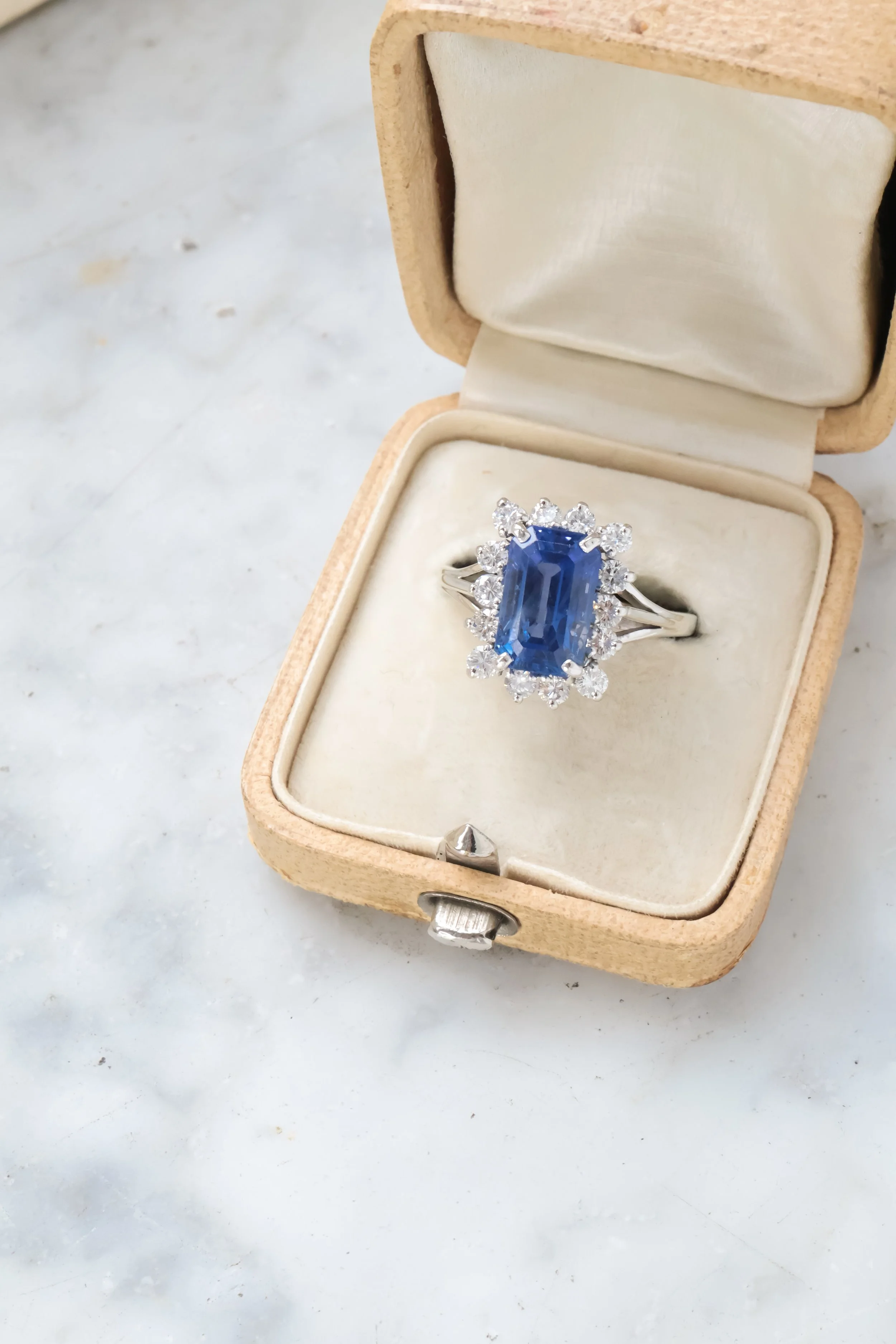 Une bague avec un gros saphir bleu entouré de diamants, présentée dans une boîte en bois avec intérieur en tissu clair.