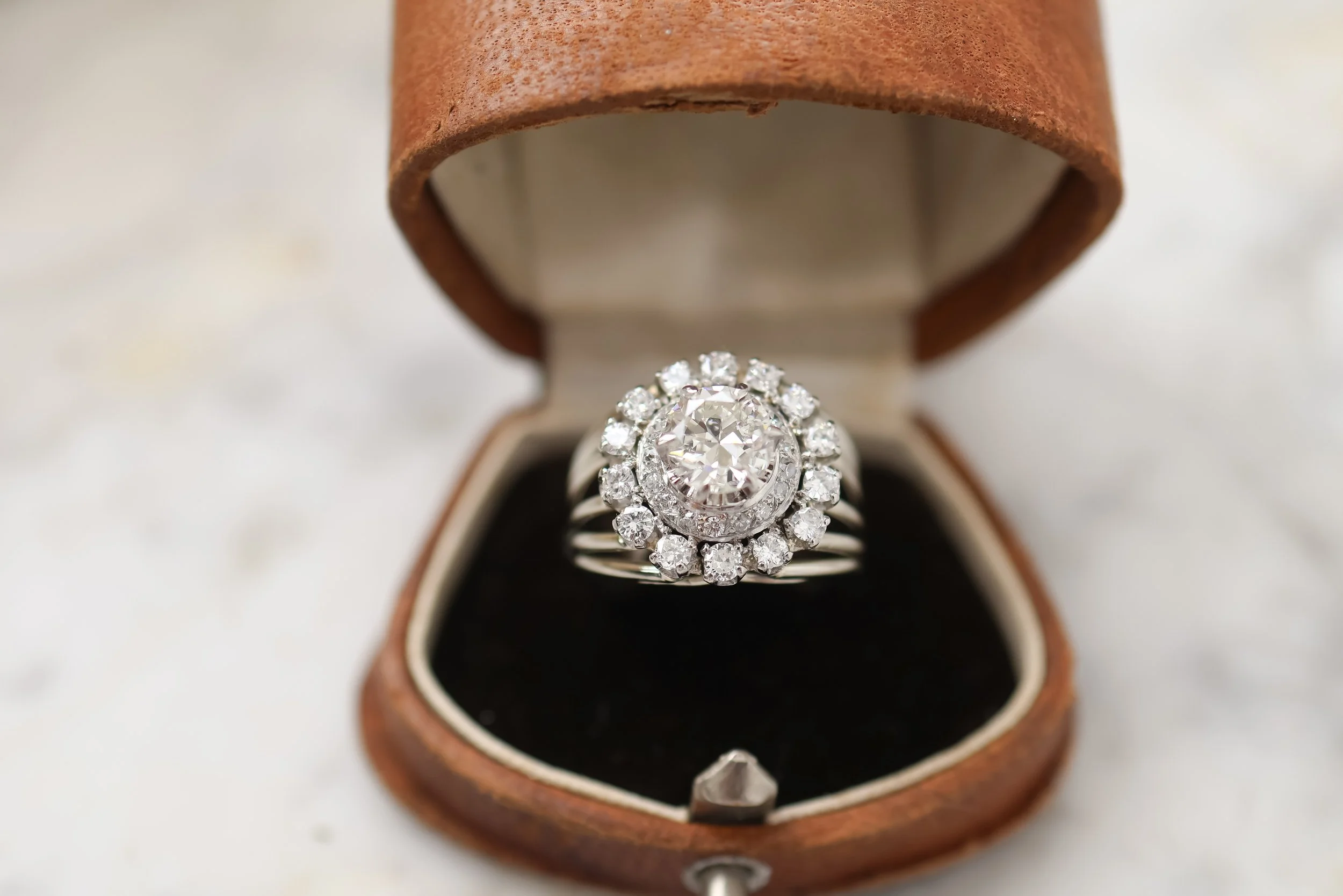 Bague en argent avec un diamant central entouré de petits diamants dans une boîte à bijoux en cuir marron.