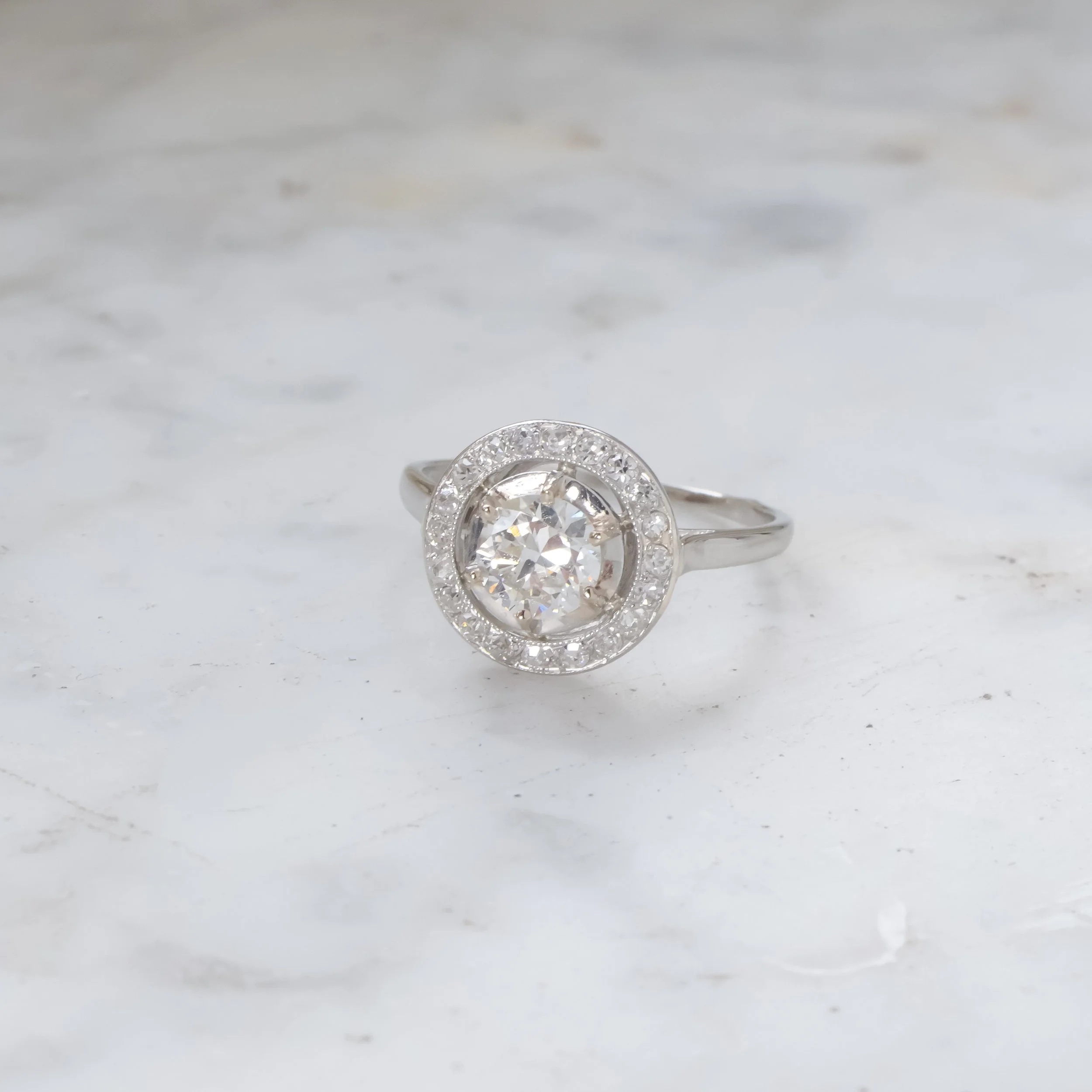 Bague en or blanc ornée d'un grand diamant central entouré de petits diamants sur une surface blanche