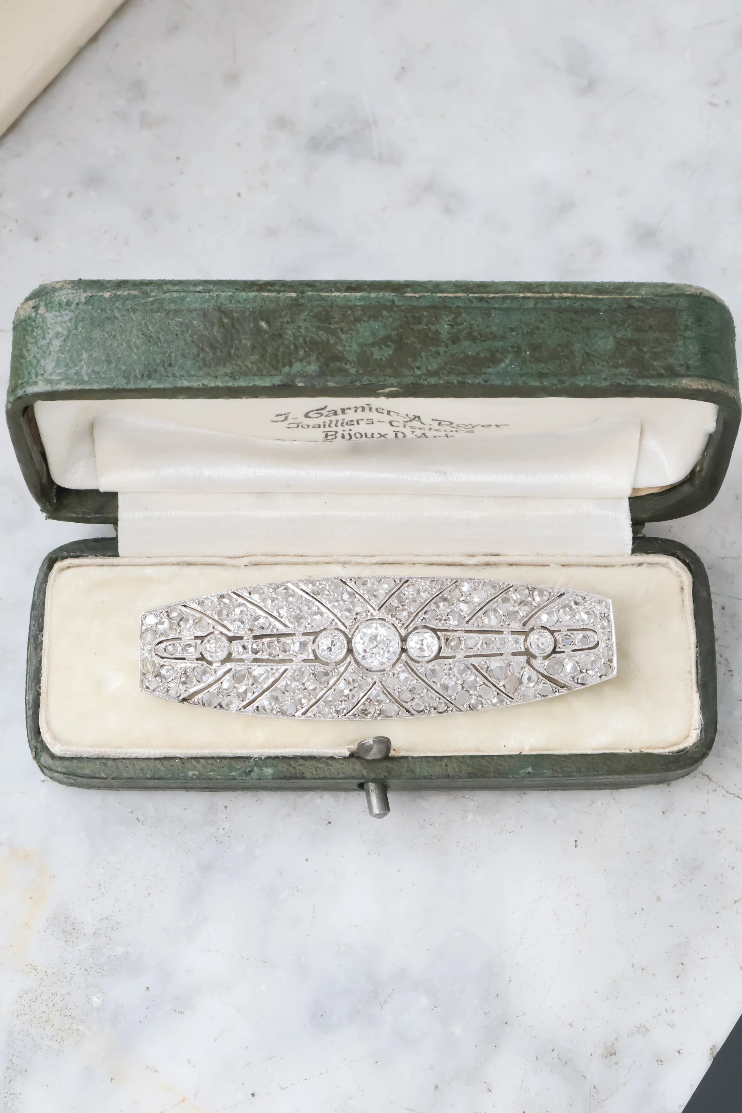 Une broche en diamants dans un écrin ancien vert, La boîte est ouverte, montrant la broche ancienne avec des motifs en diamants.