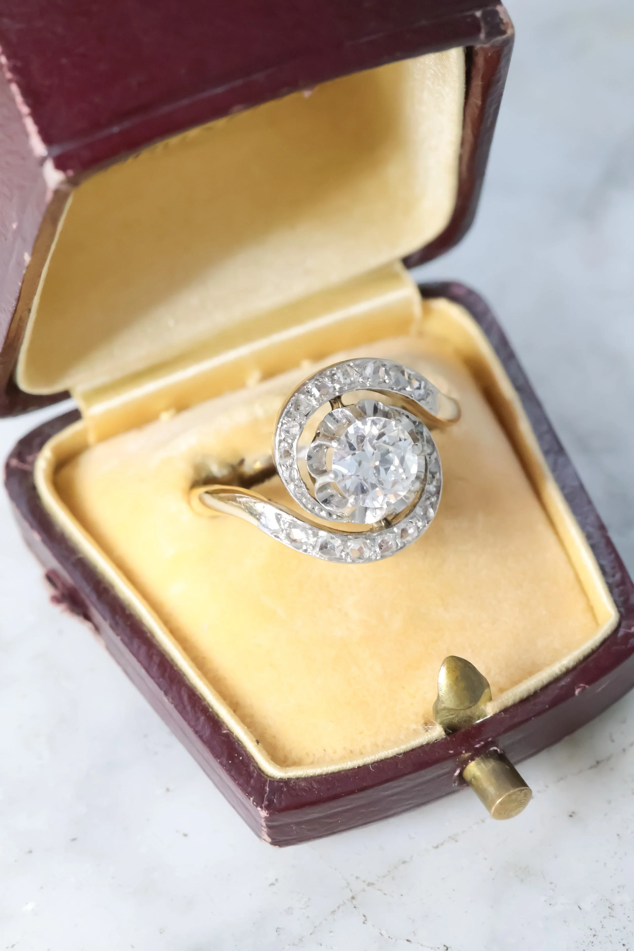 Bague ancienne tourbillon en or sertie d’un diamant central entouré de diamants, présentée dans un écrin vintage en bois