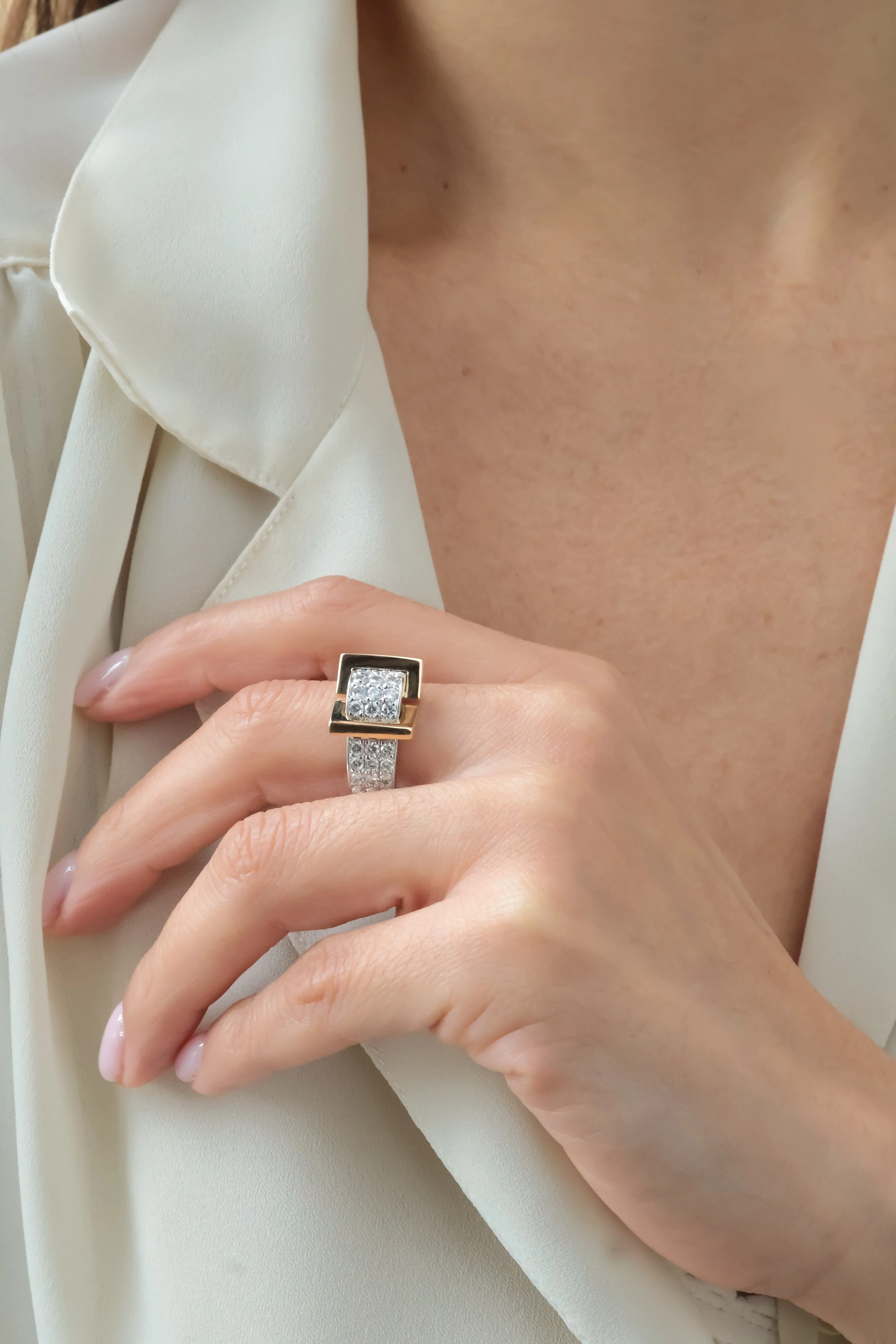 Une main portant une bague en or blanc avec des diamants, posée sur une veste blanche ou crème. La personne porte également une veste ou un chemisier blanc.