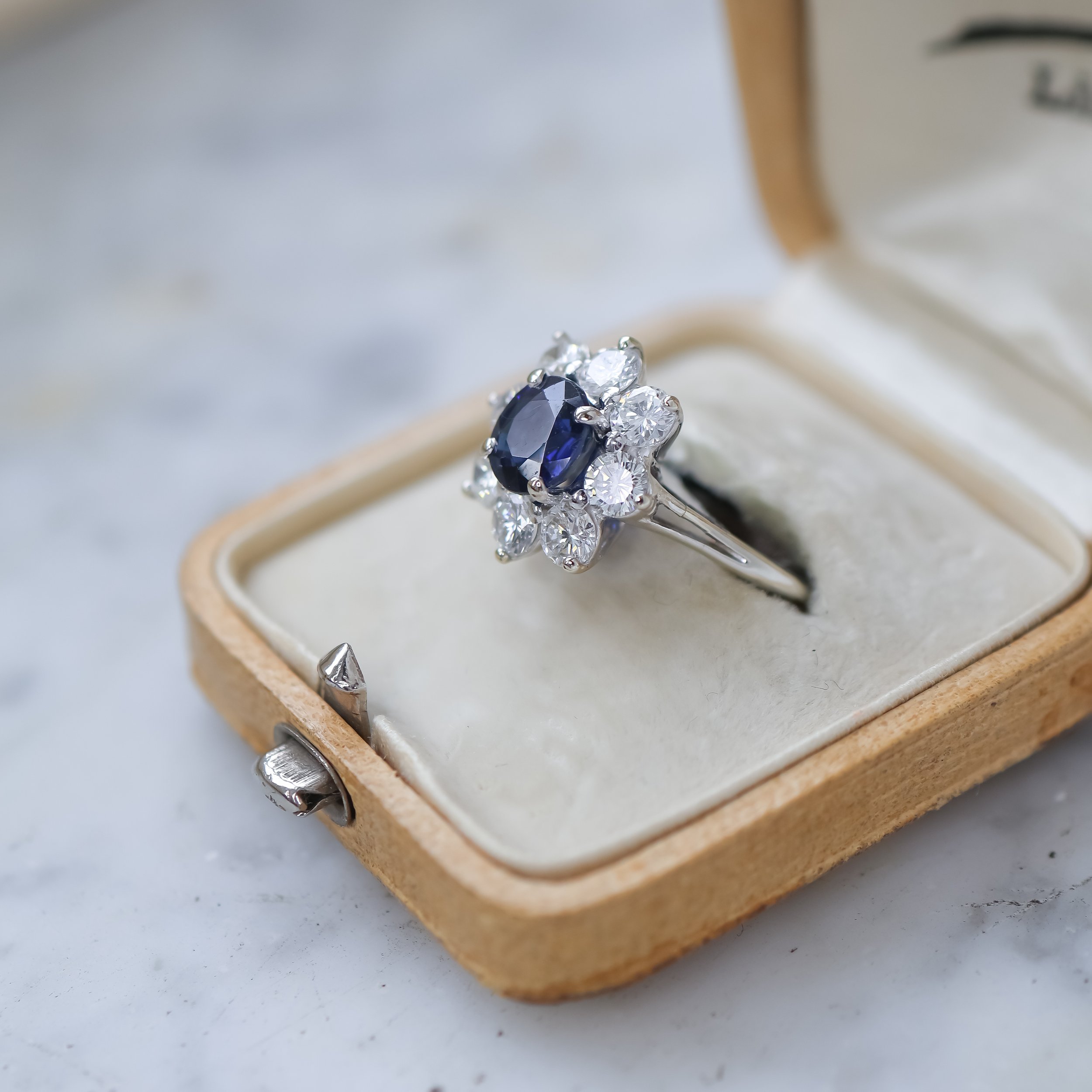 Bague en argent avec un saphir oval noir entouré de diamants, dans une boîte à bijoux en velours blanc.