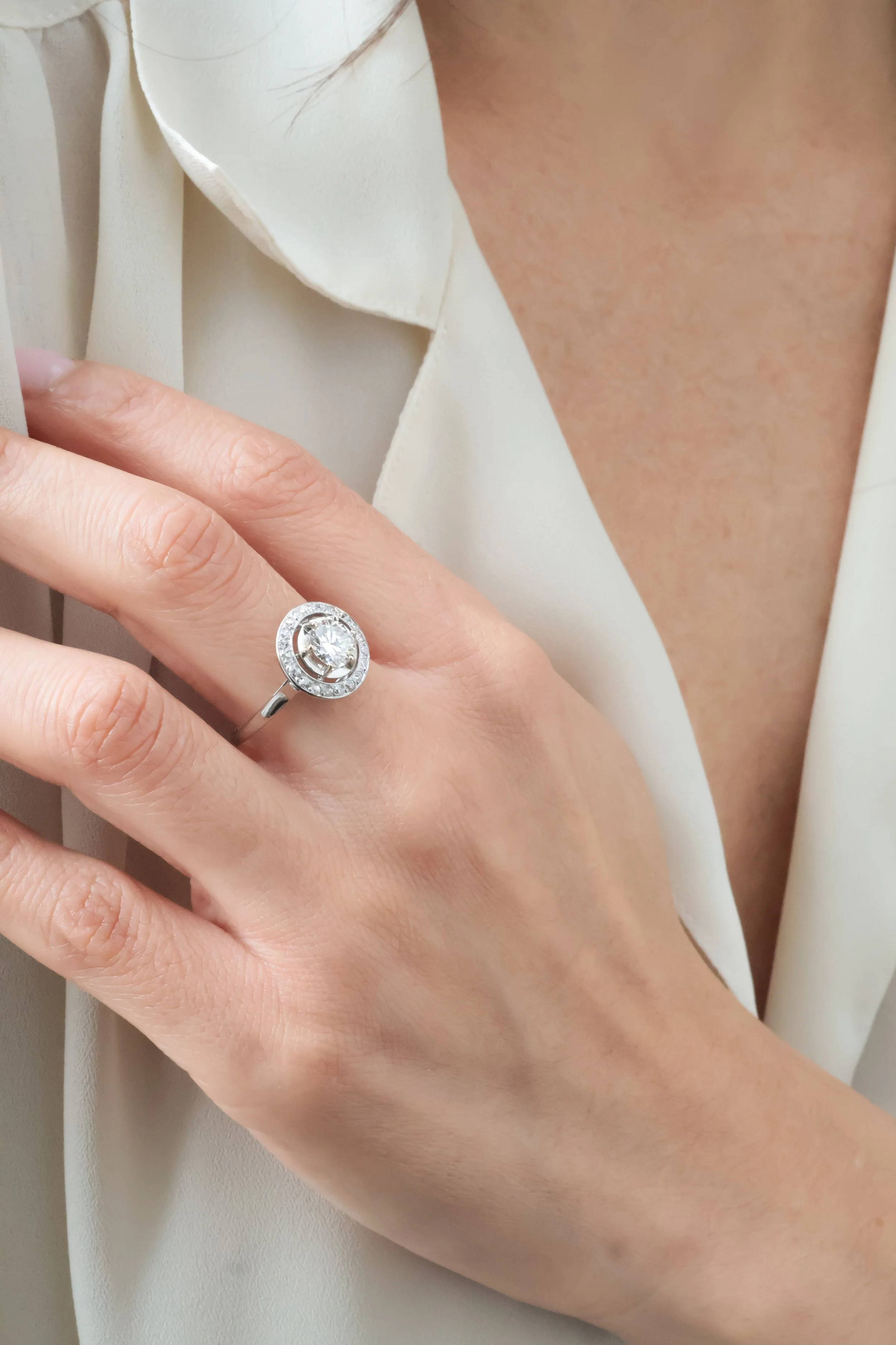 Une main portant une bague en argent avec un diamant au centre, entourée de petits diamants, et une personne portant un vêtement beige ou crème à col en V.