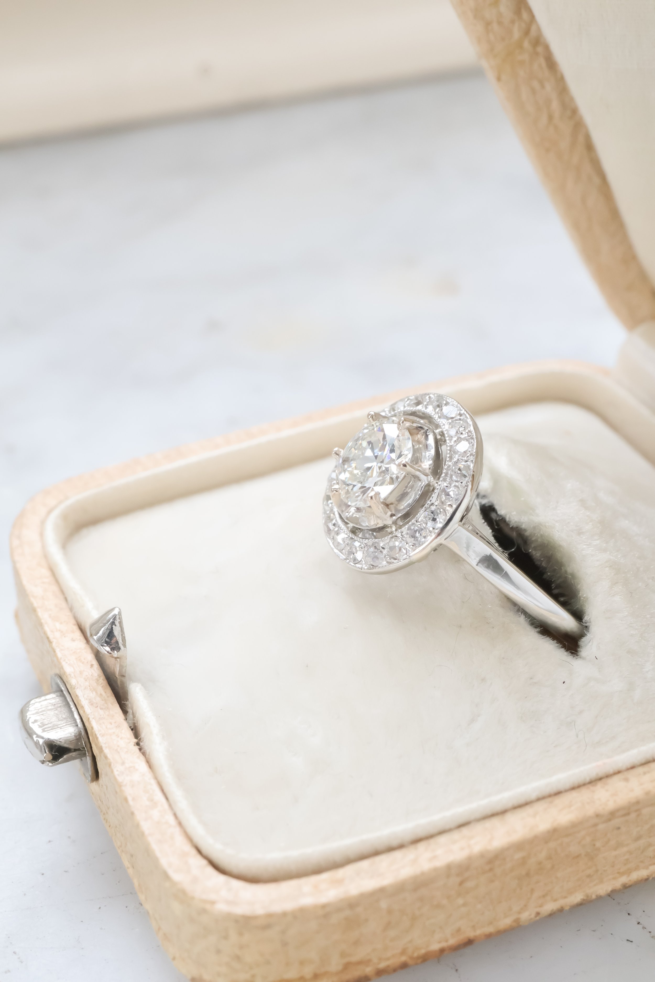 Bague en argent avec un grand diamant circulaire entouré de petits diamants, présentée dans une boîte en bois avec intérieur en tissu blanc.