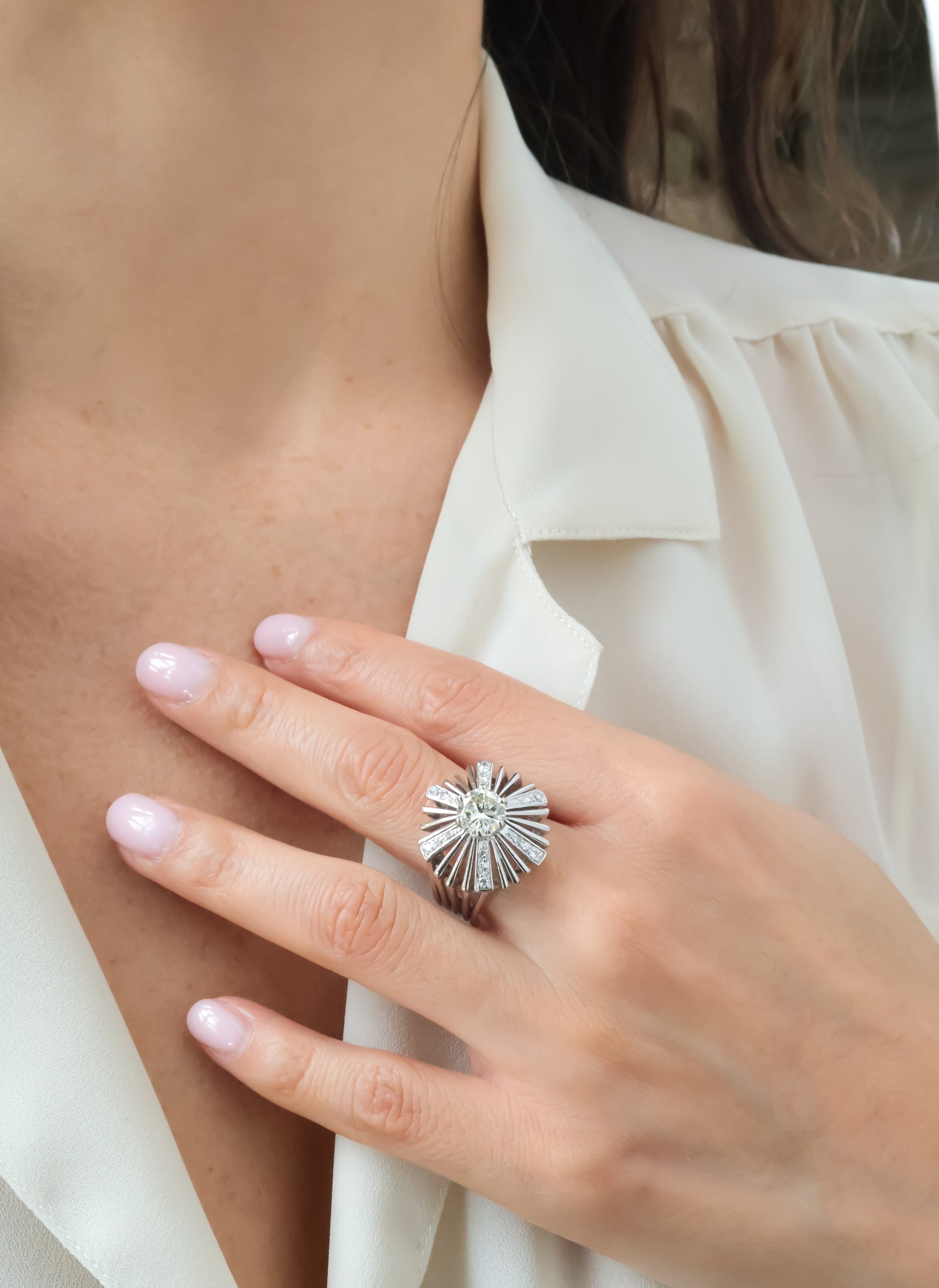 Une main portant une bague ornée d'un grand diamant au centre, avec un design rayonnant, posée sur la poitrine d'une femme en blouse blanche. La personne a la peau claire, et la blouse est en tissu léger et beige.