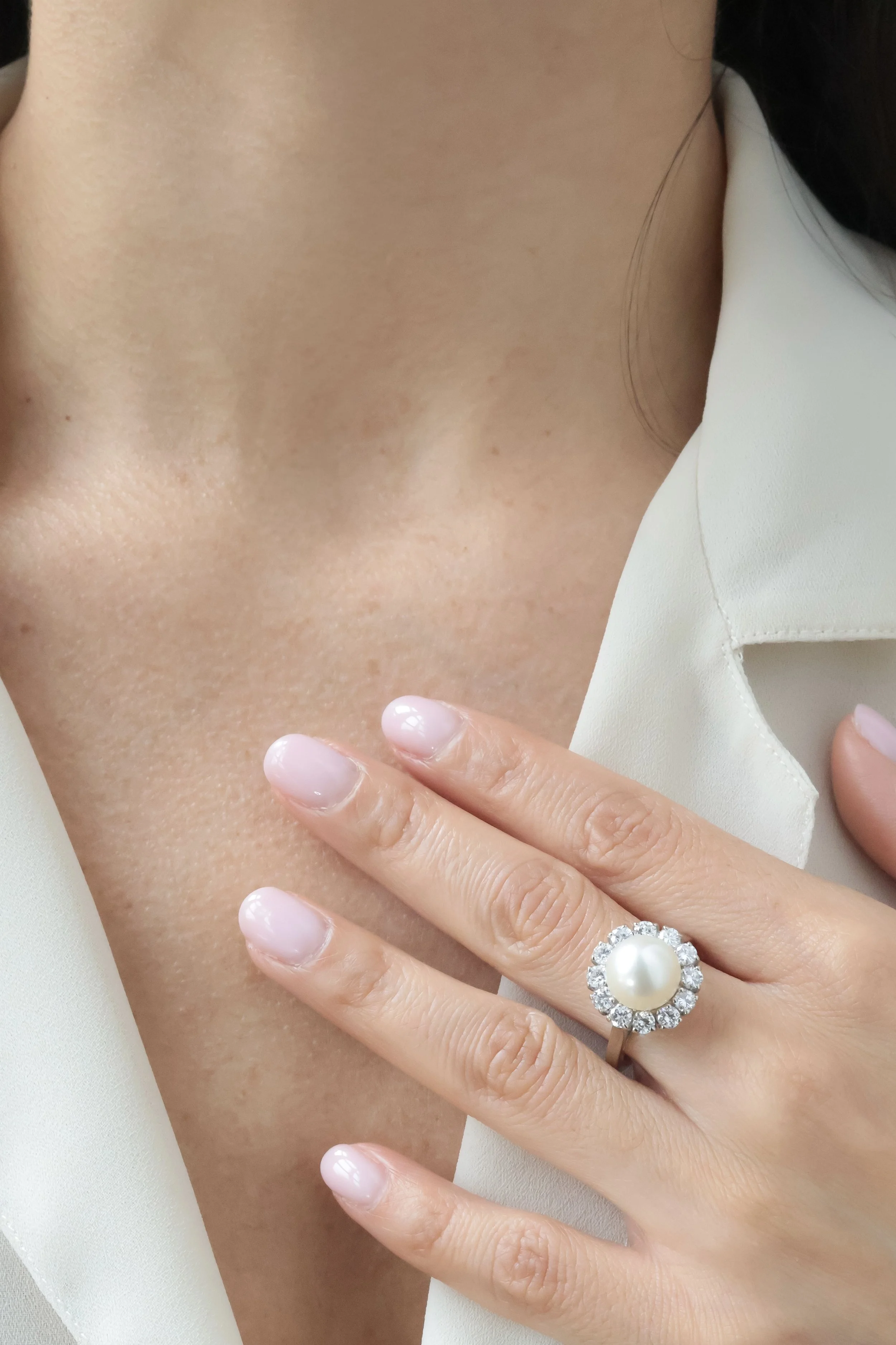 Doigt d'une femme portant une bague avec perle centrale entourée de diamants, posé sur le cou ou la poitrine nue, vêtue d'une chemise blanche.