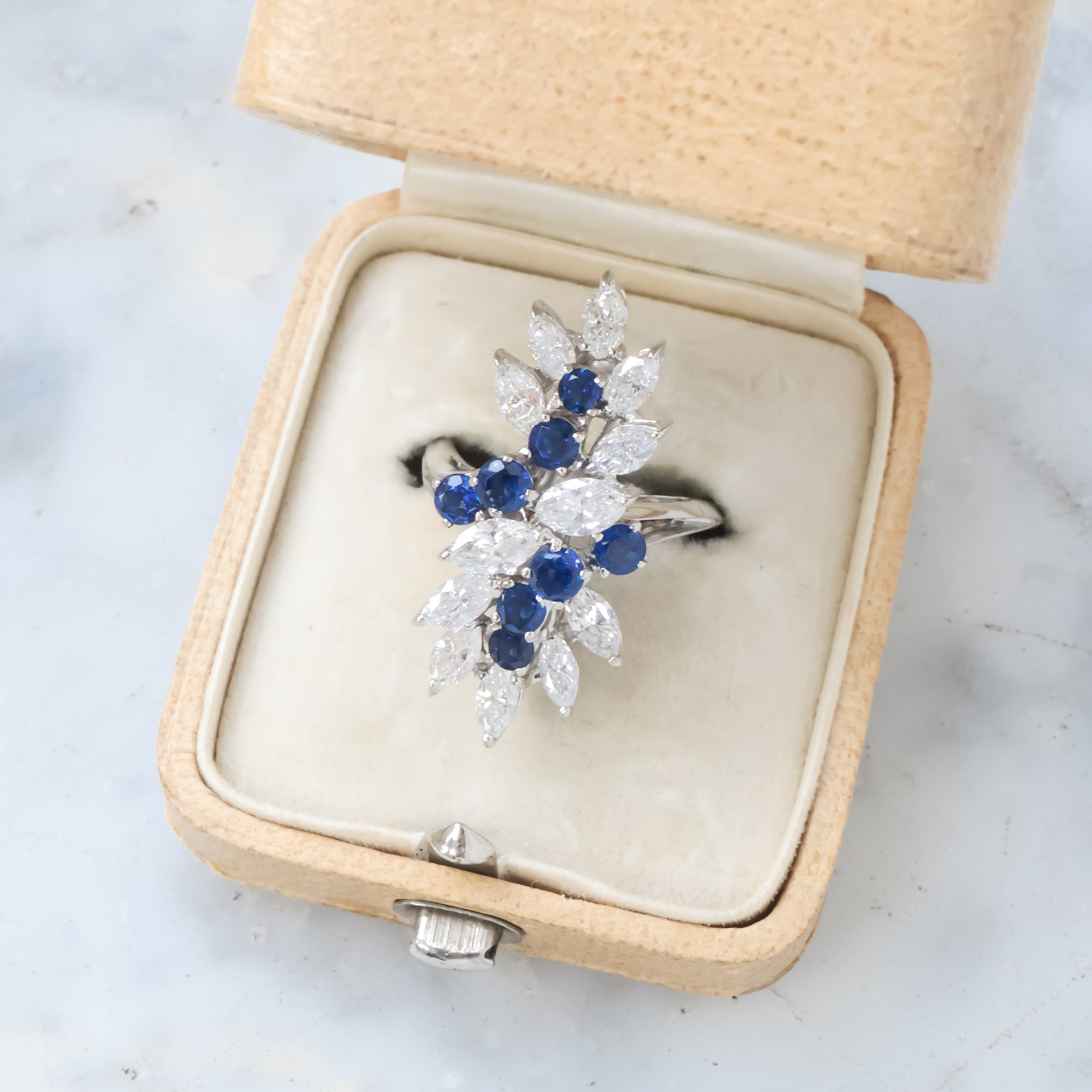 Bague cocktail en or blanc ornée de diamants taille navette et de saphirs ronds dans un écrin beige 