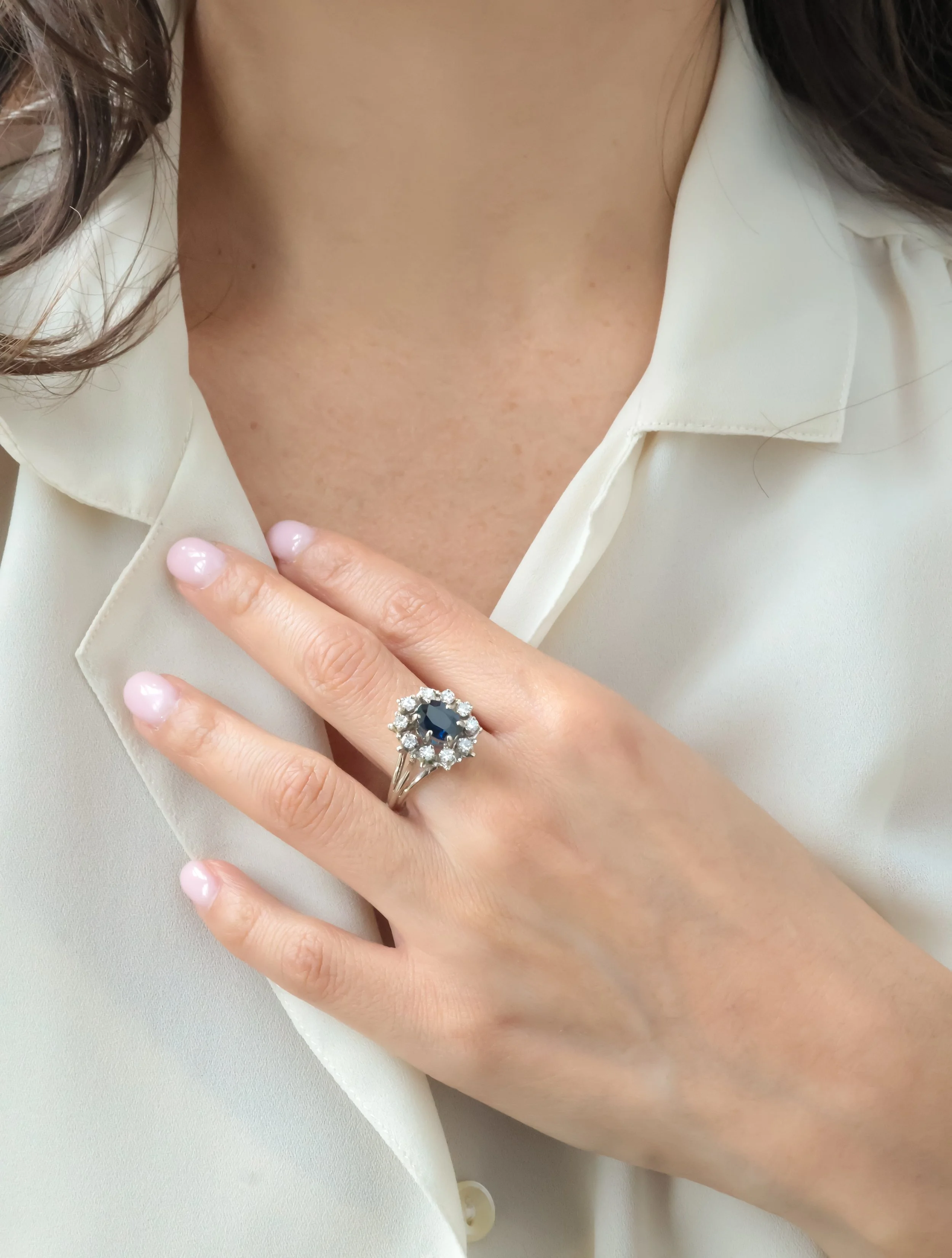 Main d'une femme portant une bague avec un grand pierre noire entourée de pierres blanches, vêtue d'une chemise claire.