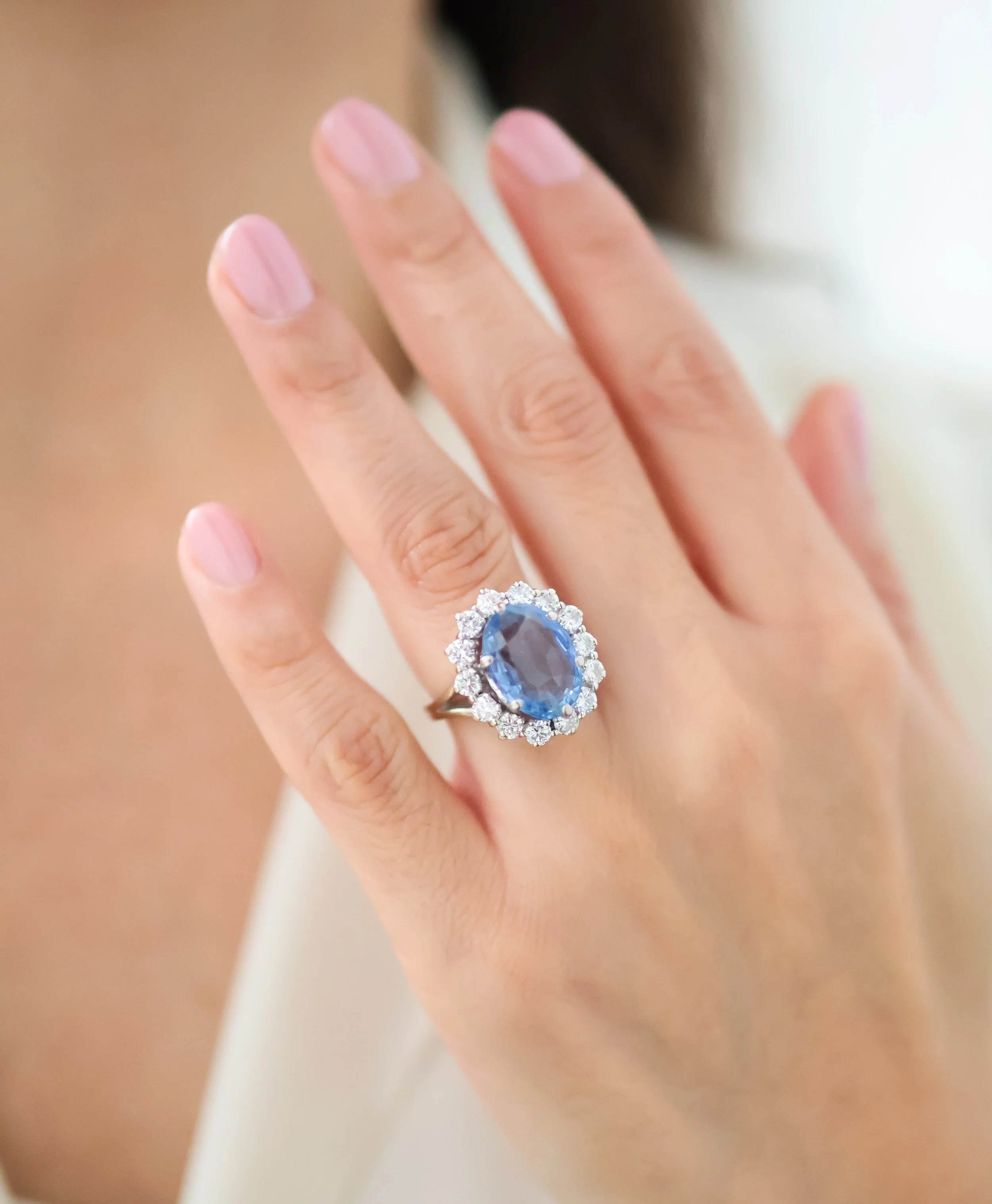 Une main portant une bague avec une pierre précieuse ovale bleue entourée de petits diamants, sur un fond neutre.