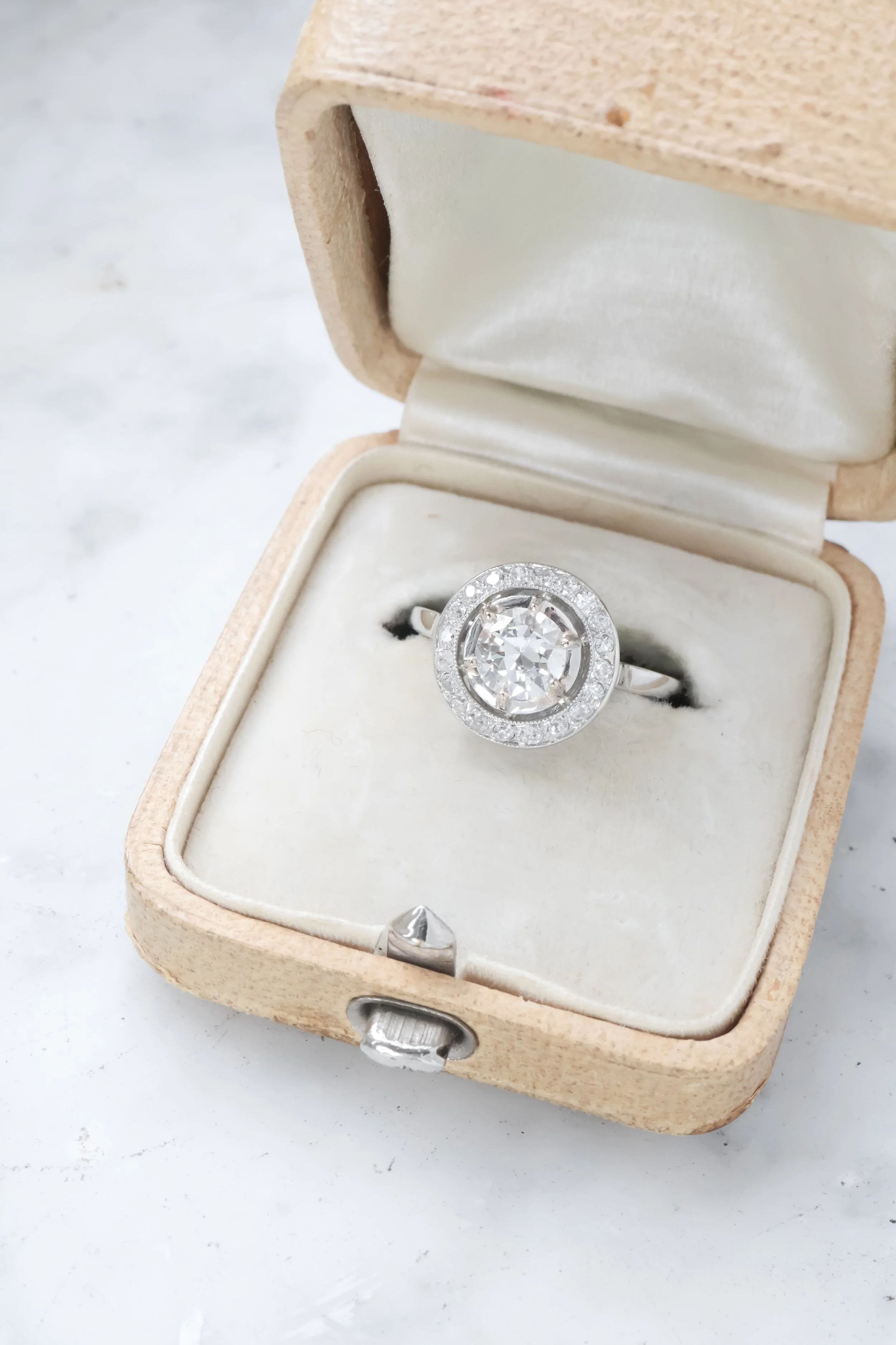 Bague en argent avec un grand diamant au centre, entourée de petits diamants, dans une boîte en bois avec intérieur en velours beige.
