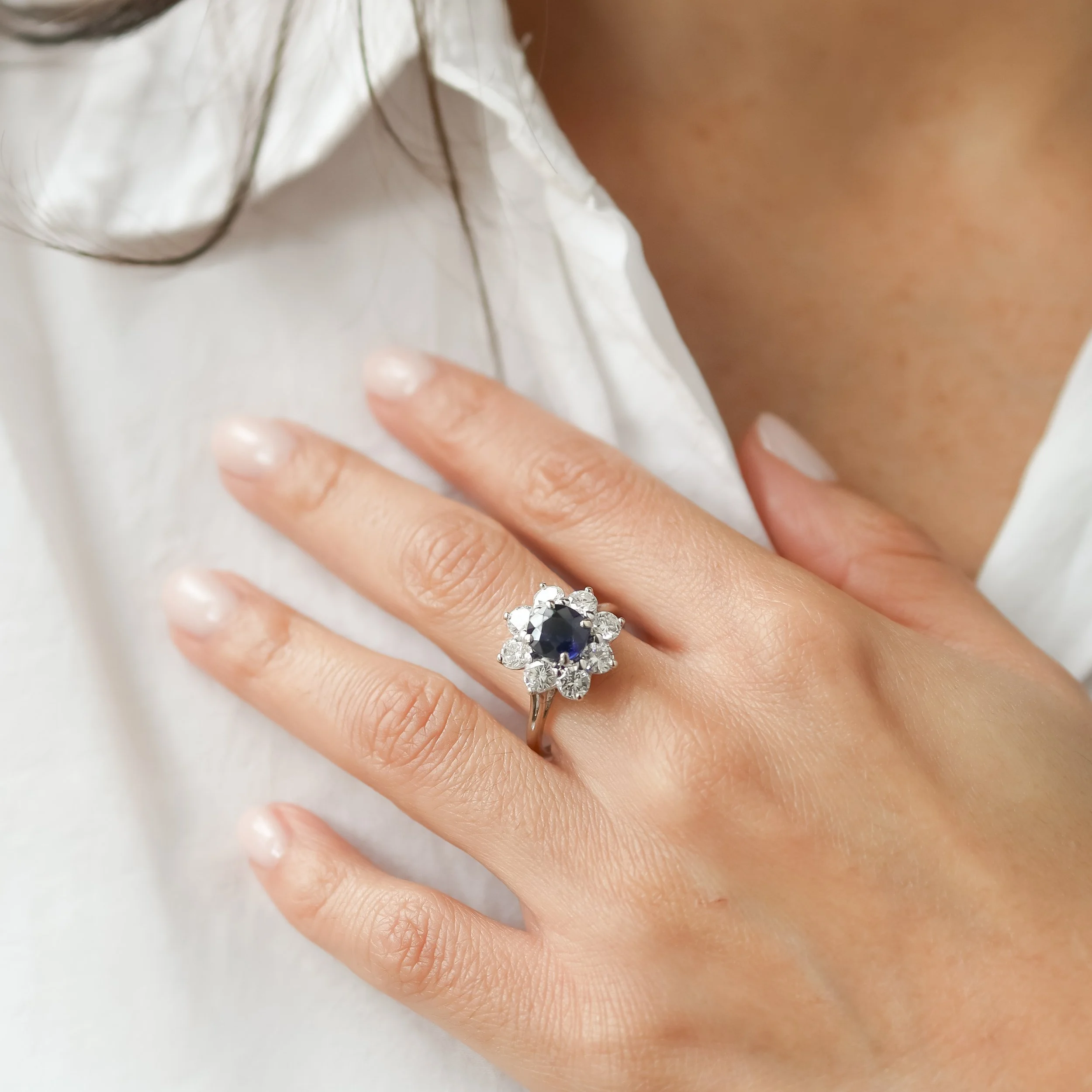 Une main portant une bague avec un saphir central entouré de diamants, posée sur une chemise blanche.
