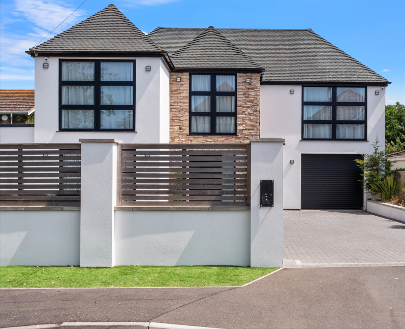 Modern two-story house with white walls, brick accents, large black-framed windows, a sloped roof, and a black garage door, surrounded by a white wall with horizontal wooden fencing.