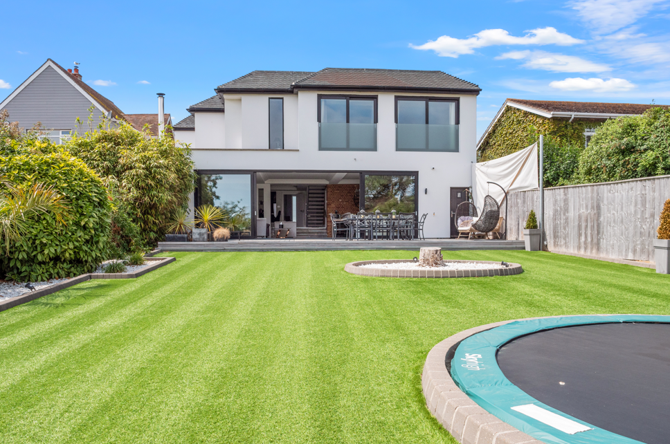 Backyard with green lawn, modern two-story house, patio furniture, trampoline, and tree and shrubbery on sides, under blue sky with a few clouds.