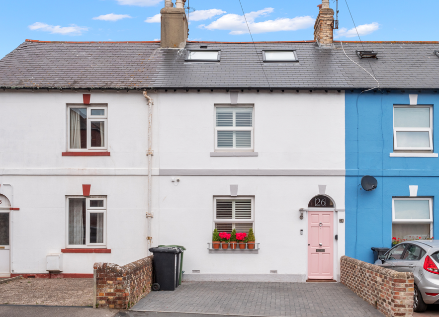 Row of colorful houses, with the middle one painted white, featuring a pink front door and flower boxes with pink flowers, and the house on the right painted blue, with a black satellite dish and a parked car in front.