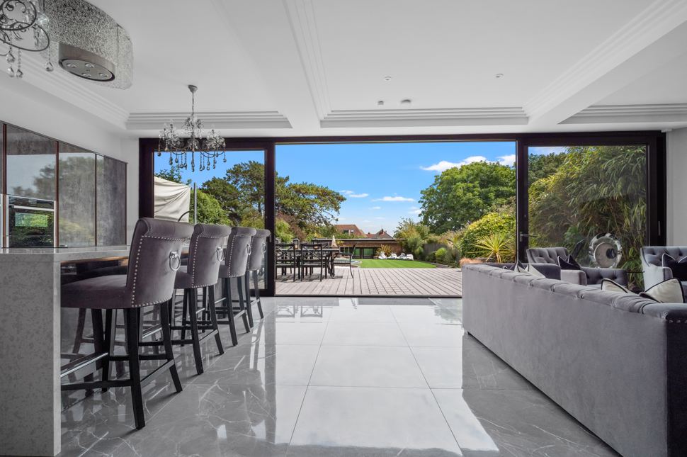 Modern living room with floor-to-ceiling glass doors leading to a patio and backyard with trees and blue sky.