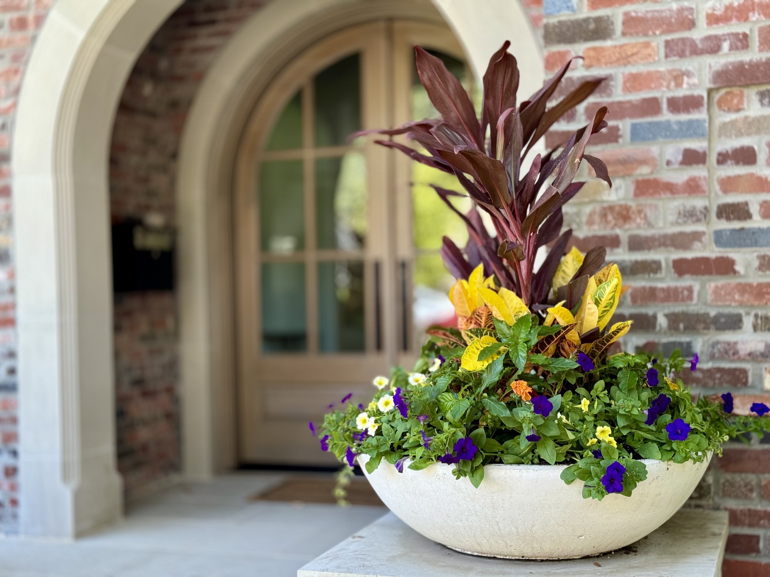 Colorful flower arrangement in an off-white bowl on a table in front of a brick wall and arched doorway.