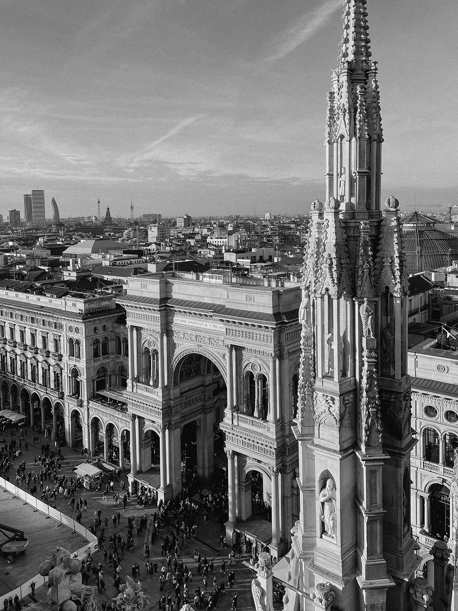 Vista dall'alto di una piazza affollata con archi e un edificio storico, con un alto pinnacolo decorato con statue.