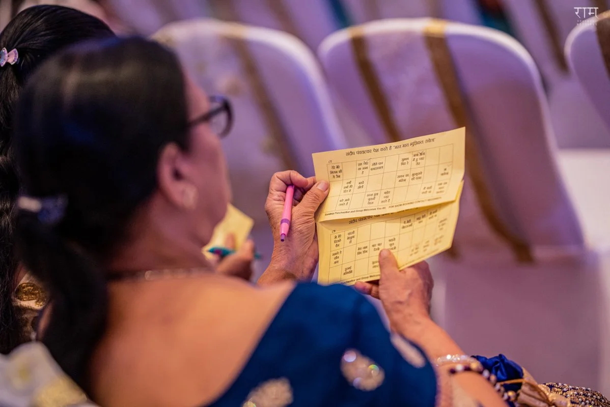 A woman with glasses and a blue saree sitting in an auditorium, holding and writing on a yellow examination or answer sheet with a pink pen. India and Canada