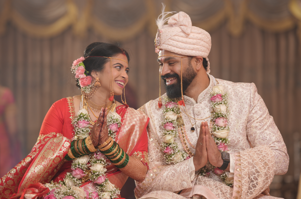 Indian bride and groom smiling with hands pressed together in a greeting gesture, dressed in traditional wedding attire with floral garlands, in a decorated ceremonial setting. India and Canada