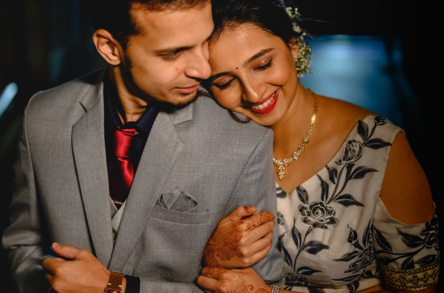 A happy couple at a celebration, with the woman showing mehndi on her hand, smiling with eyes closed, leaning on each other, dressed elegantly. India and Canada