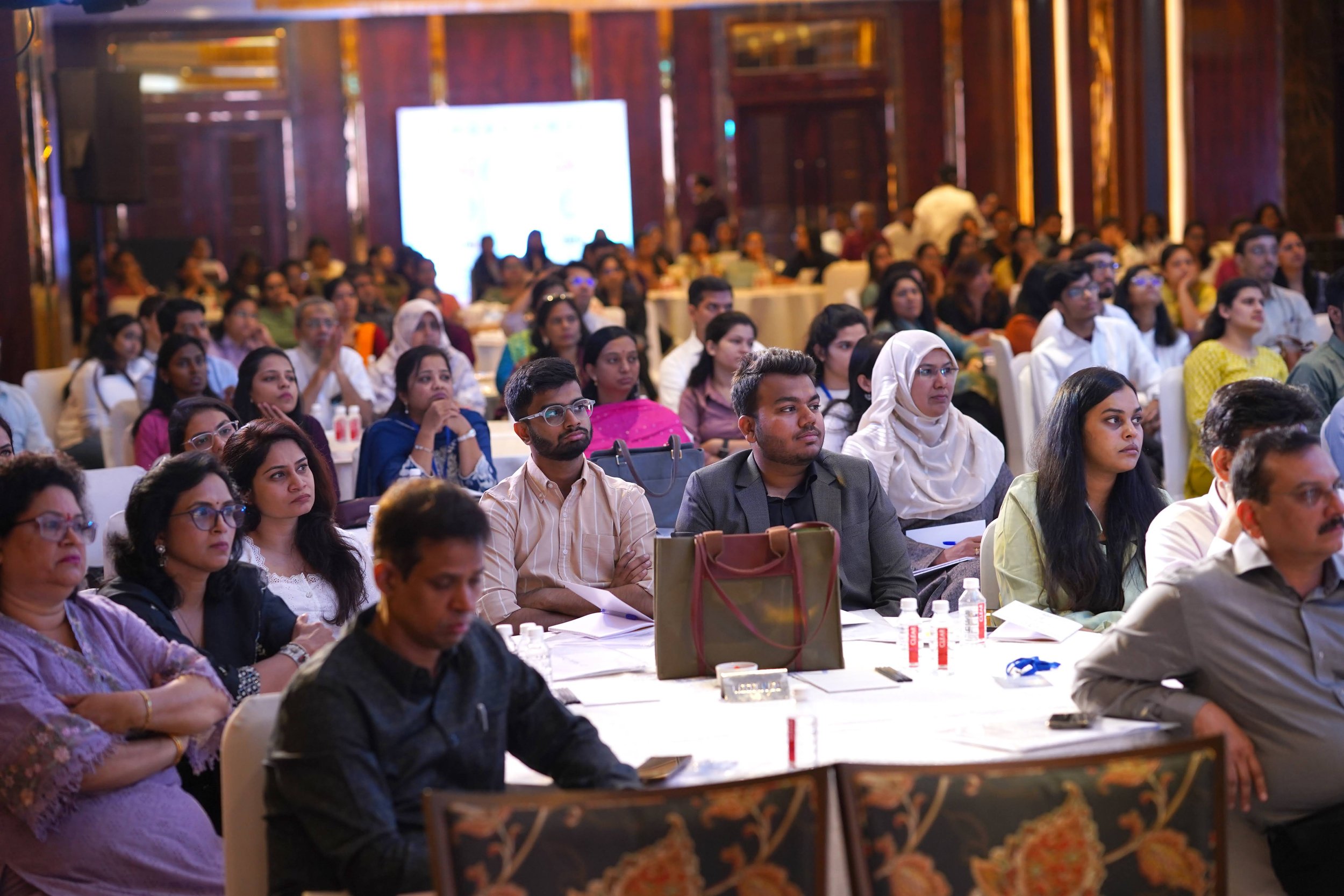 Attendees listening attentively at a conference in a large, well-lit room with wooden paneling, sitting at round tables with water bottles and notebooks. India and Canada