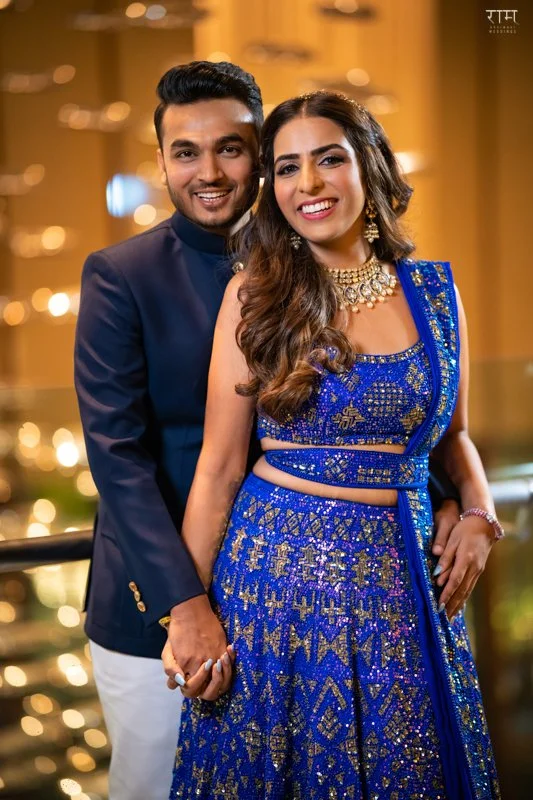 A smiling couple A man and a woman standing close together, holding hands, dressed in traditional Indian clothing, with the woman wearing a blue embroidered lehenga and jewelry, and the man wearing a navy blue kurta, at a festive event with warm bokeh lighting in the background.